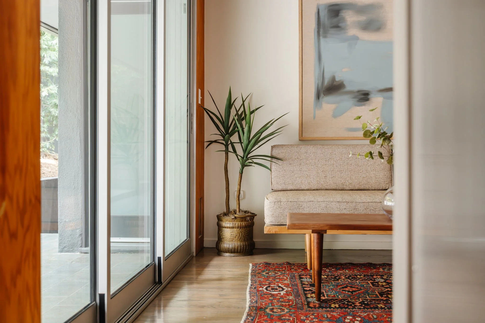  Vignette of a neutral MCM daybed on top of a red and blue Persian rug. A small floor plant in a brass planter sits to the left of the daybed, with beige and blue abstract artwork hung above. 
