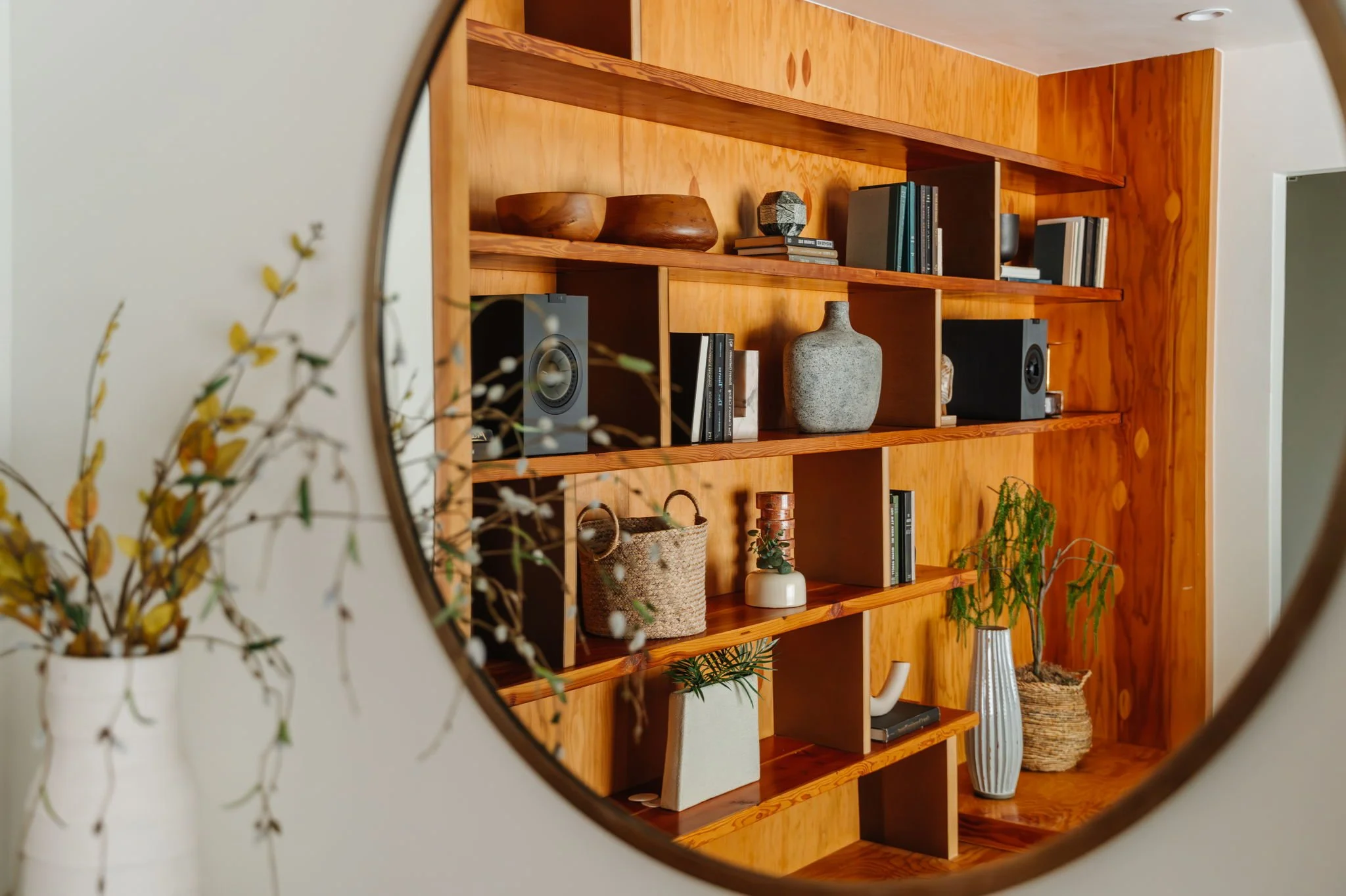  Vignette of a round mirror reflecting a styled solid wood built-in bookcase. The shelves are decorated with books and found objects.  