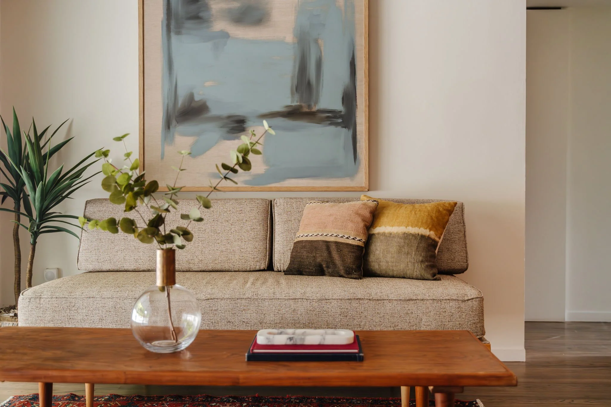  Vignette of a neutral MCM daybed on top of a red and blue Persian rug. A small floor plant in a brass planter sits to the left of the daybed, with beige and blue abstract artwork hung above. 