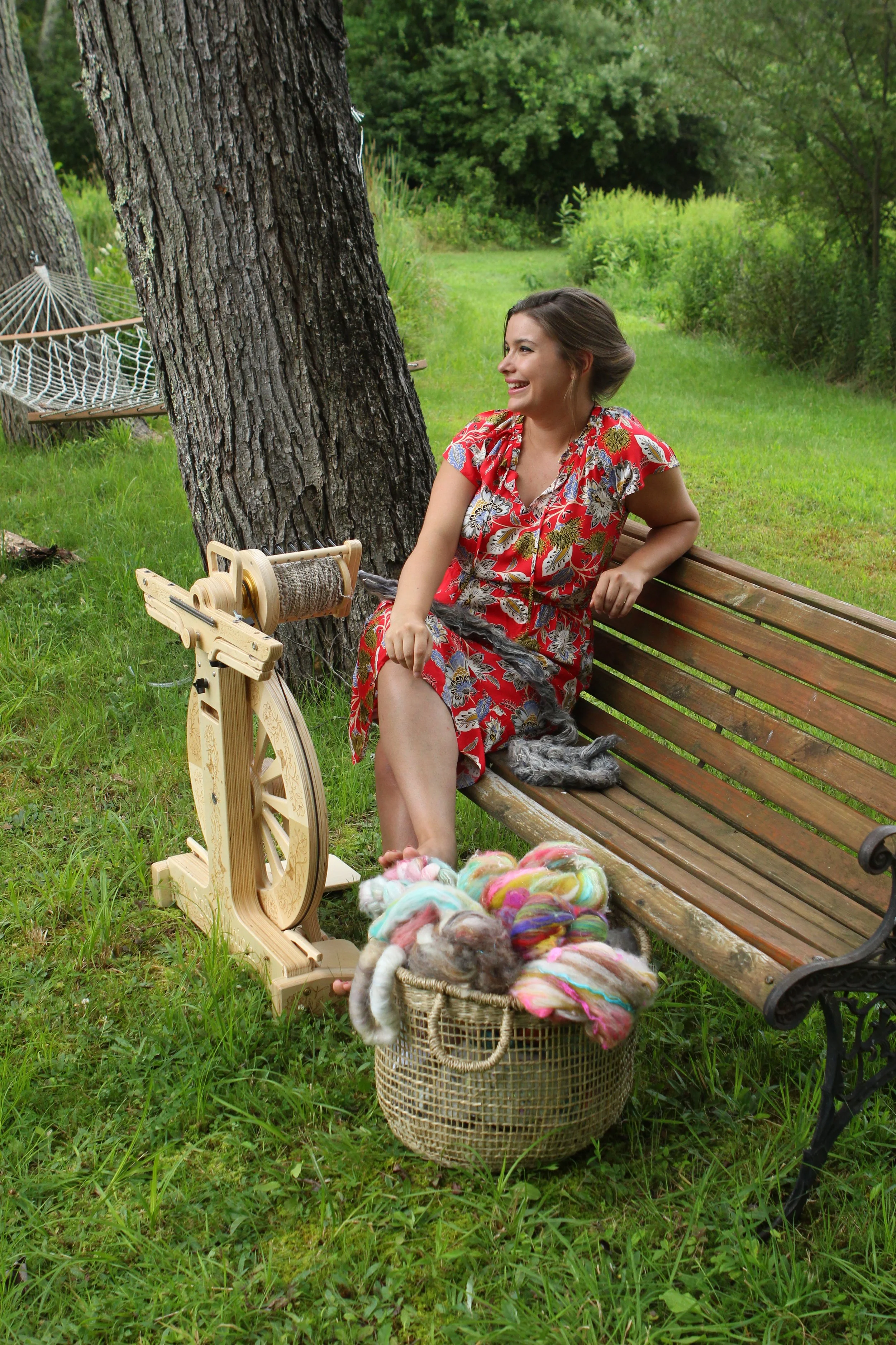 A woman in a red floral dress sitting on a wooden park bench next to a spinning wheel, surrounded by green grass and trees, with a basket of colorful wool next to her, smiling and enjoying the outdoor setting.