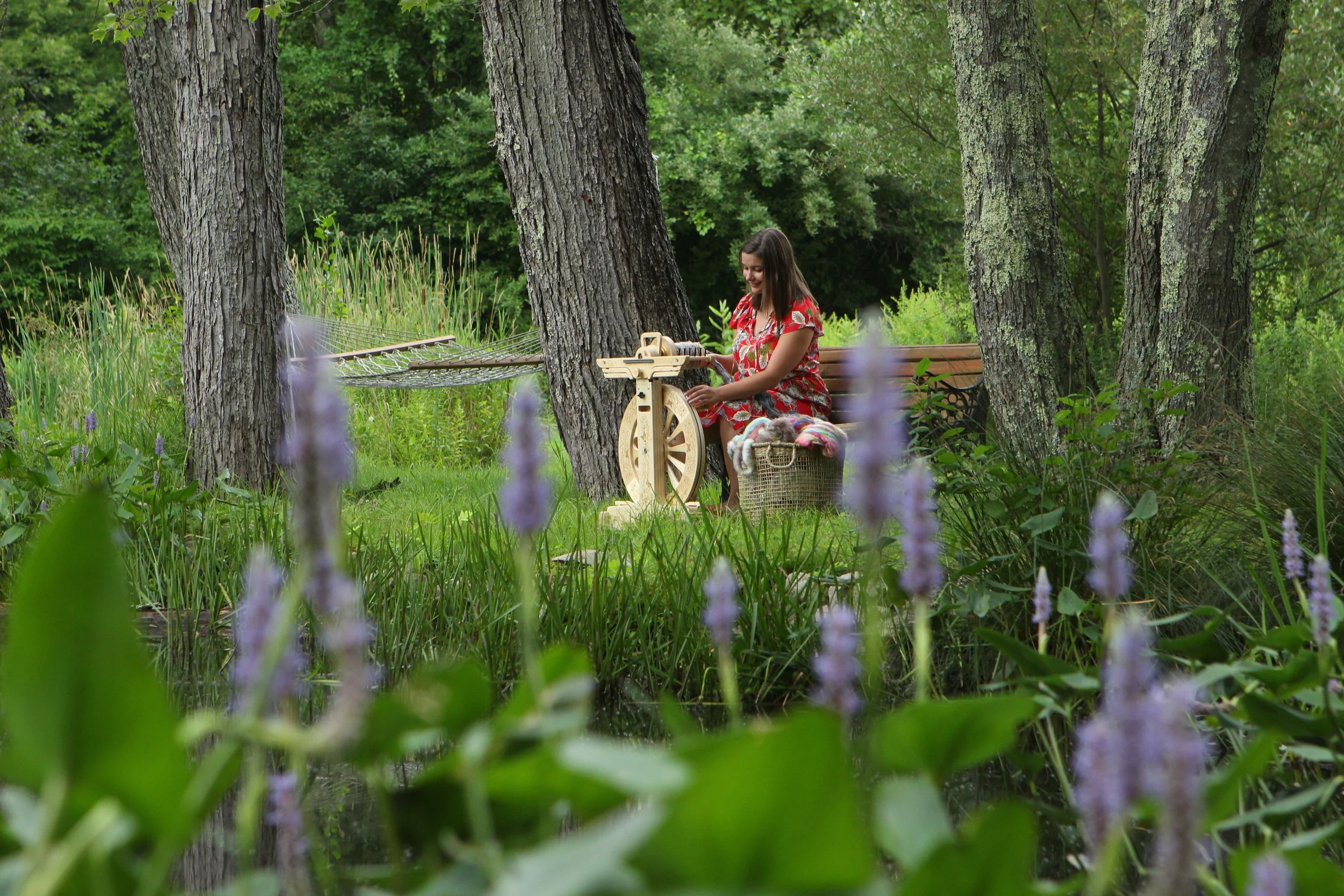 A woman in a red floral dress sitting on a wooden bench by a pond in a wooded area, operating a vintage spinning wheel, with a basket of yarn nearby.