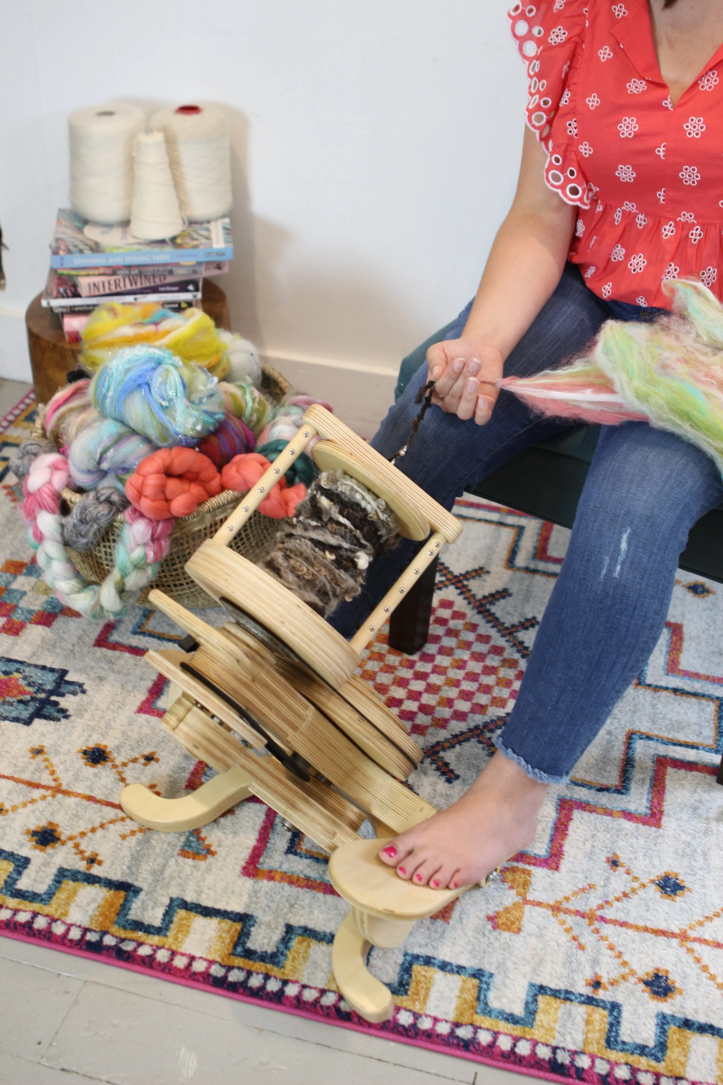 Person sitting on a chair spinning wool yarn on a terraced wooden yarn winder, with colorful wool yarns and fibers nearby, against a white wall and a patterned area rug.