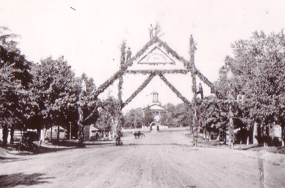 This photograph probably shows a celebration of a fire department achievement or event. The words in the top triangle are “Hook and Ladder No. 1.” There is a man standing atop each post. The old courthouse is visible at the north end of Main Street. Courtesy of Milne Library