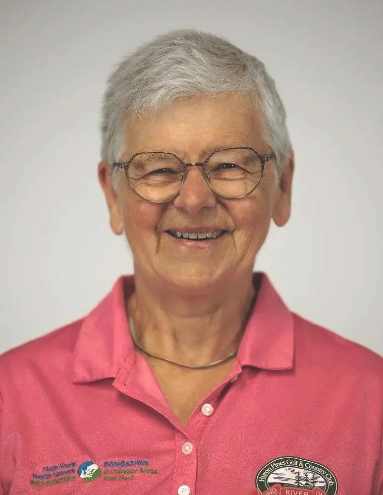 An elderly person with short gray hair and glasses, wearing a lavender and gray sweater, smiling in front of a wooden background.
