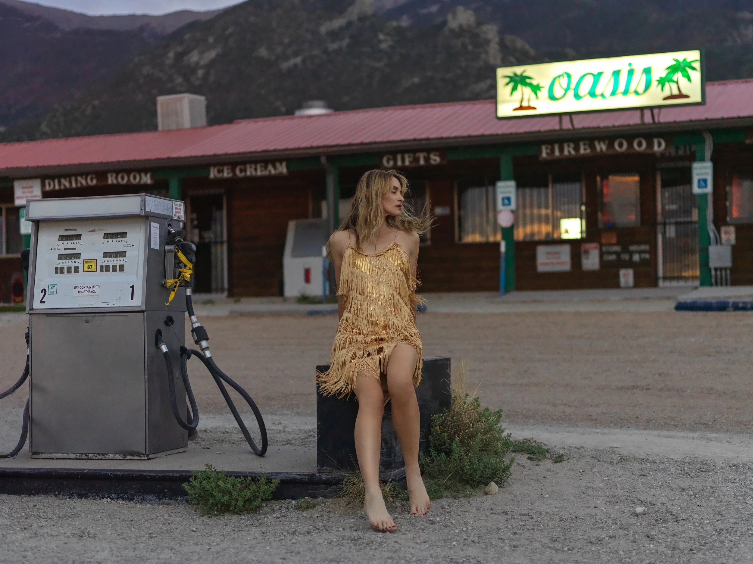A woman in a gold fringed dress standing barefoot near a gas pump at a rustic roadside stop with a sign for 'Oasis' featuring palm trees, a mountain backdrop, and a wooden building with signs for dining room, ice cream, gifts, and firewood.