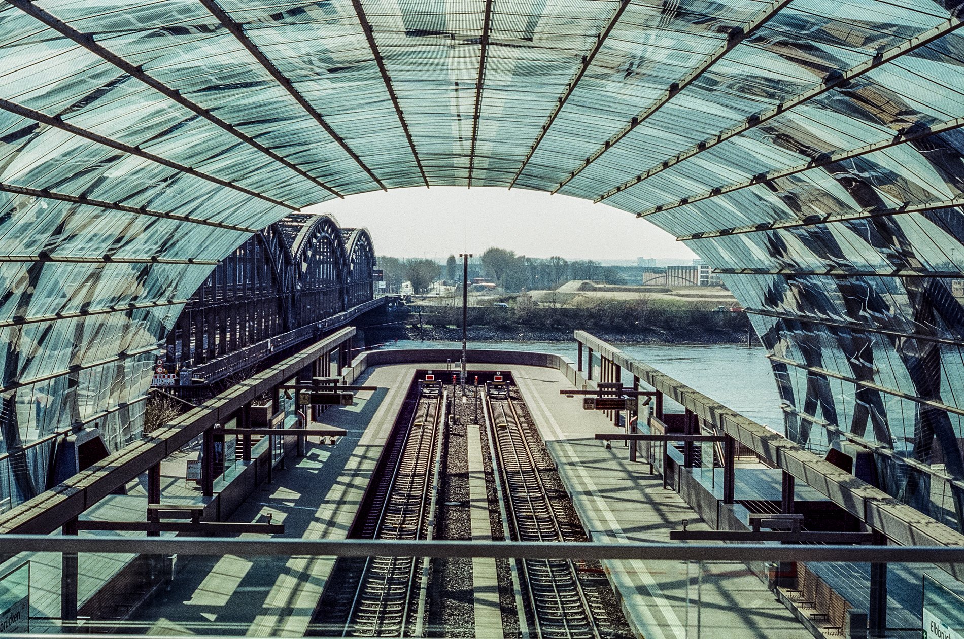 View from a train platform under a modern glass canopy, showing train tracks leading to a bridge, with water and landscape in the background.