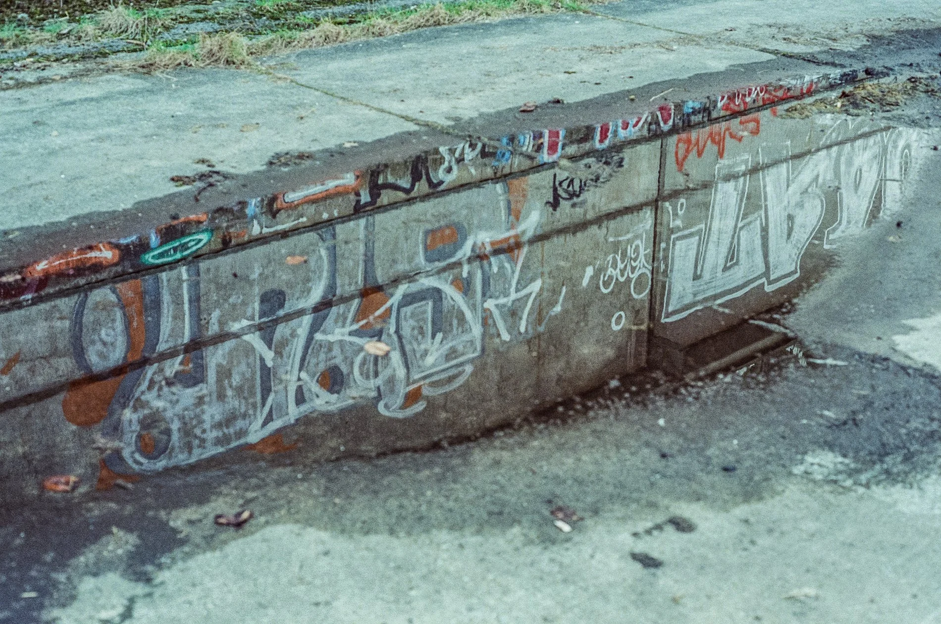 A concrete sidewalk curb covered in colorful graffiti and street art. The curb has a chipped edge and is next to a paved street surface. The graffiti includes various tags, symbols, and designs in multiple colors.