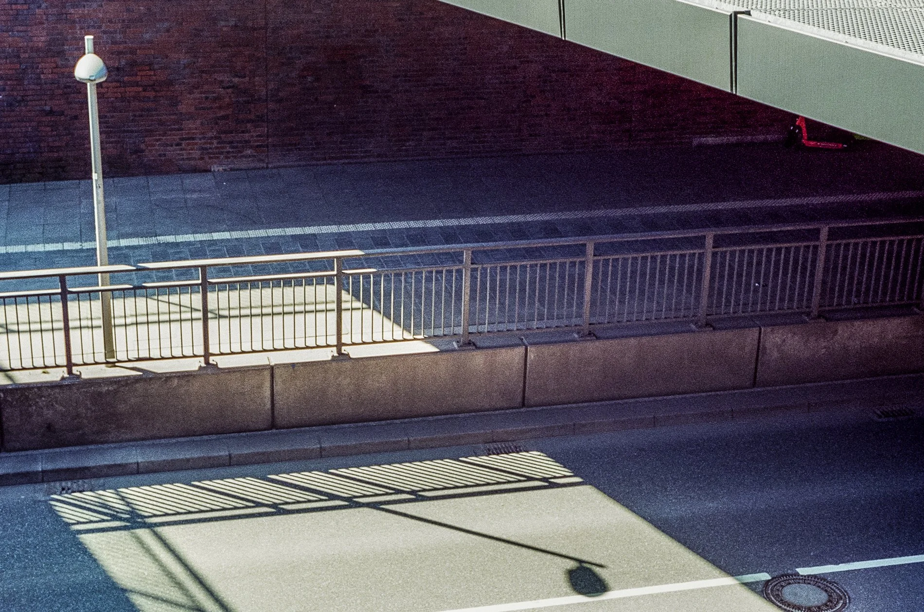 A dark street scene with a metal fence, a streetlamp, and shadows cast by urban elements. There is a brick wall in the background and a green structure overhead.