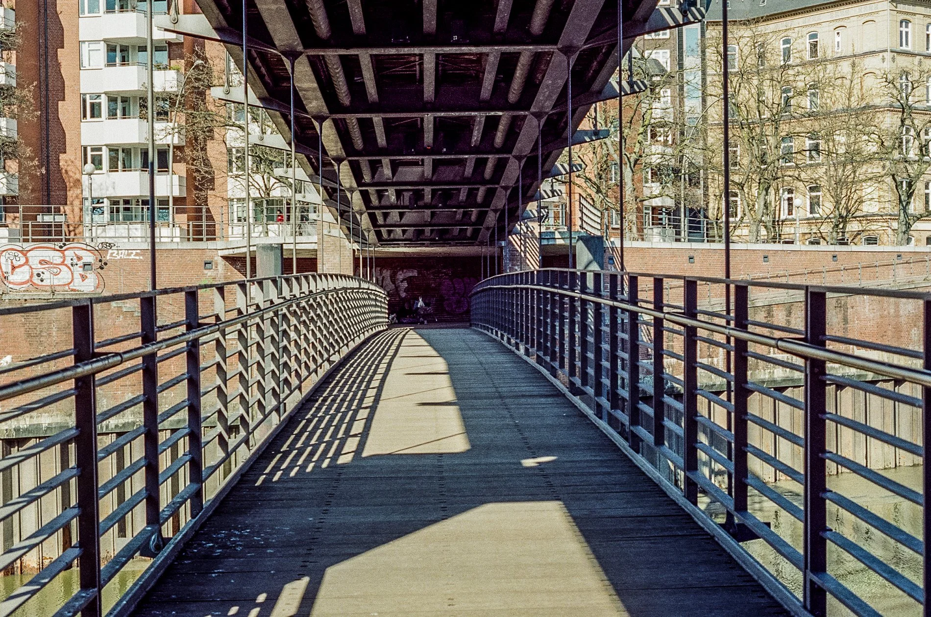 A metal pedestrian bridge with railings over a waterway in an urban area, with residential buildings and leafless trees in the background, casting shadows on the walkway.