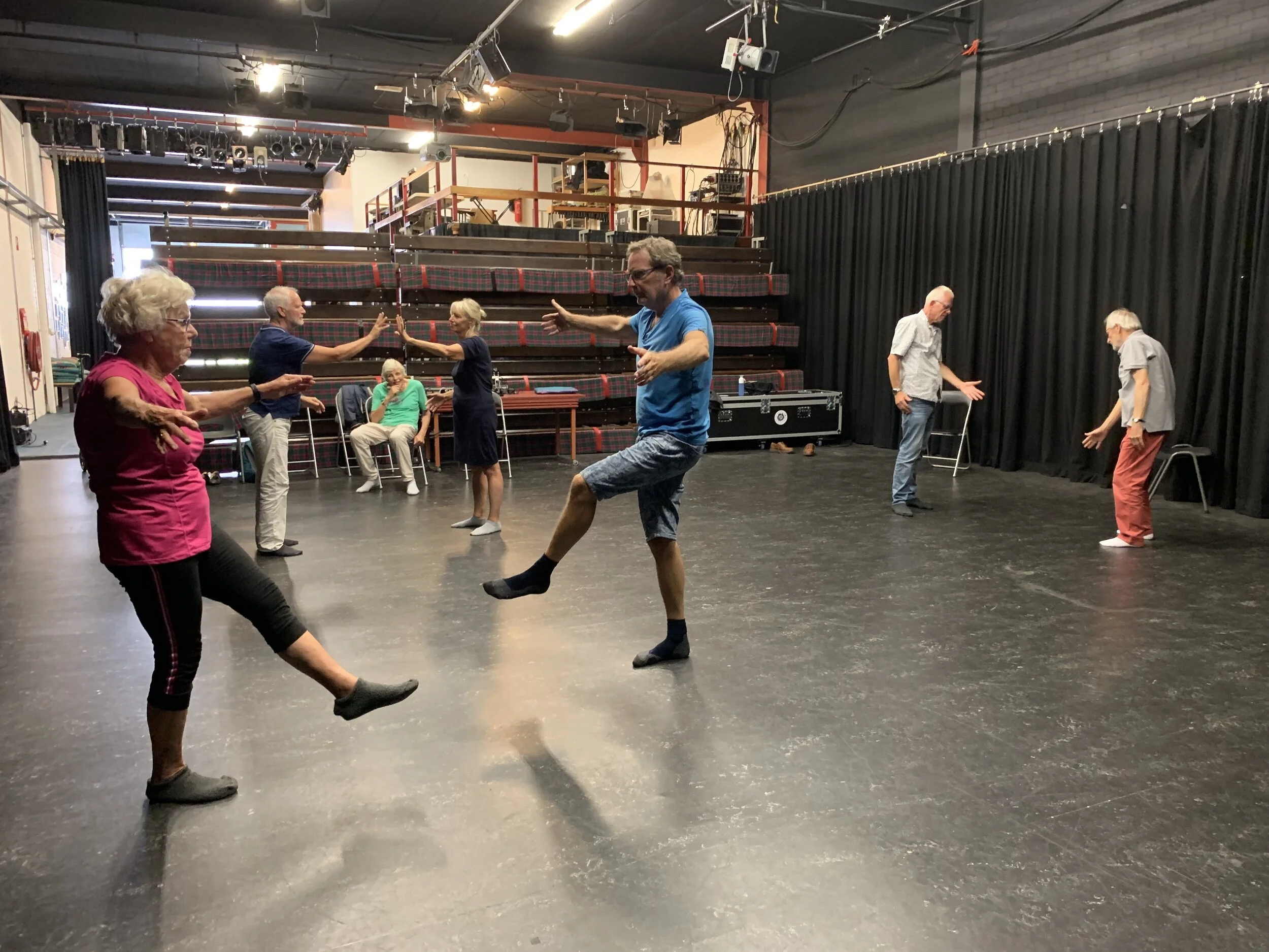 Groep mensen die samen dansen op een podium in een theater of oefenruimte, met een zwarte gordijn op de achtergrond en technische apparatuur aan het plafond.