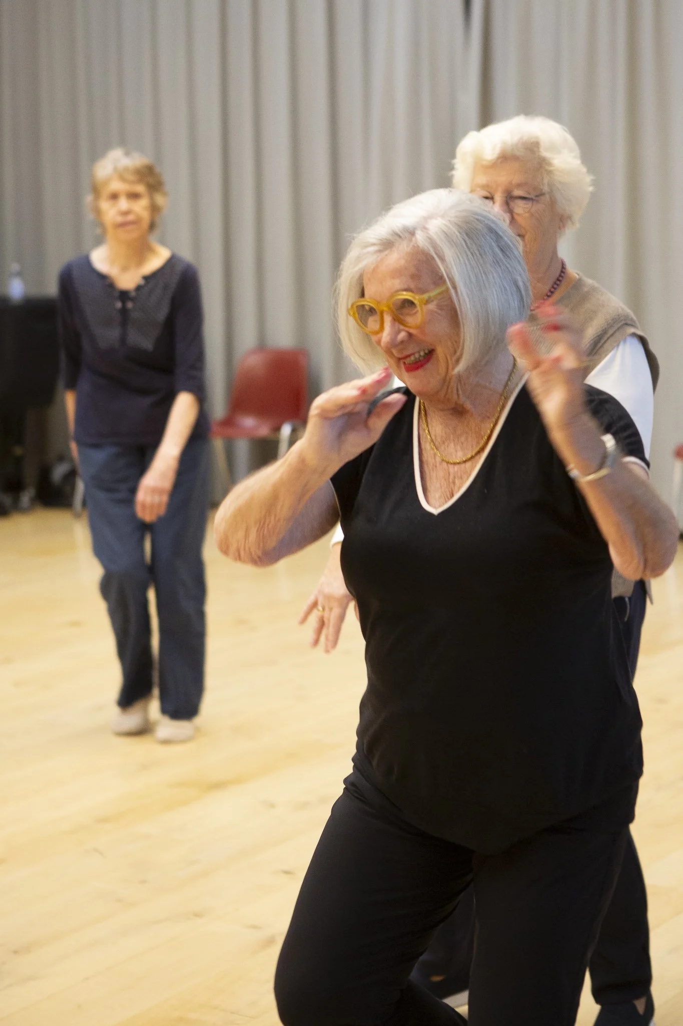 Vijf ouderen dansen samen in een zaal met houten vloer en witte gordijnen op de achtergrond, lachen en genieten.