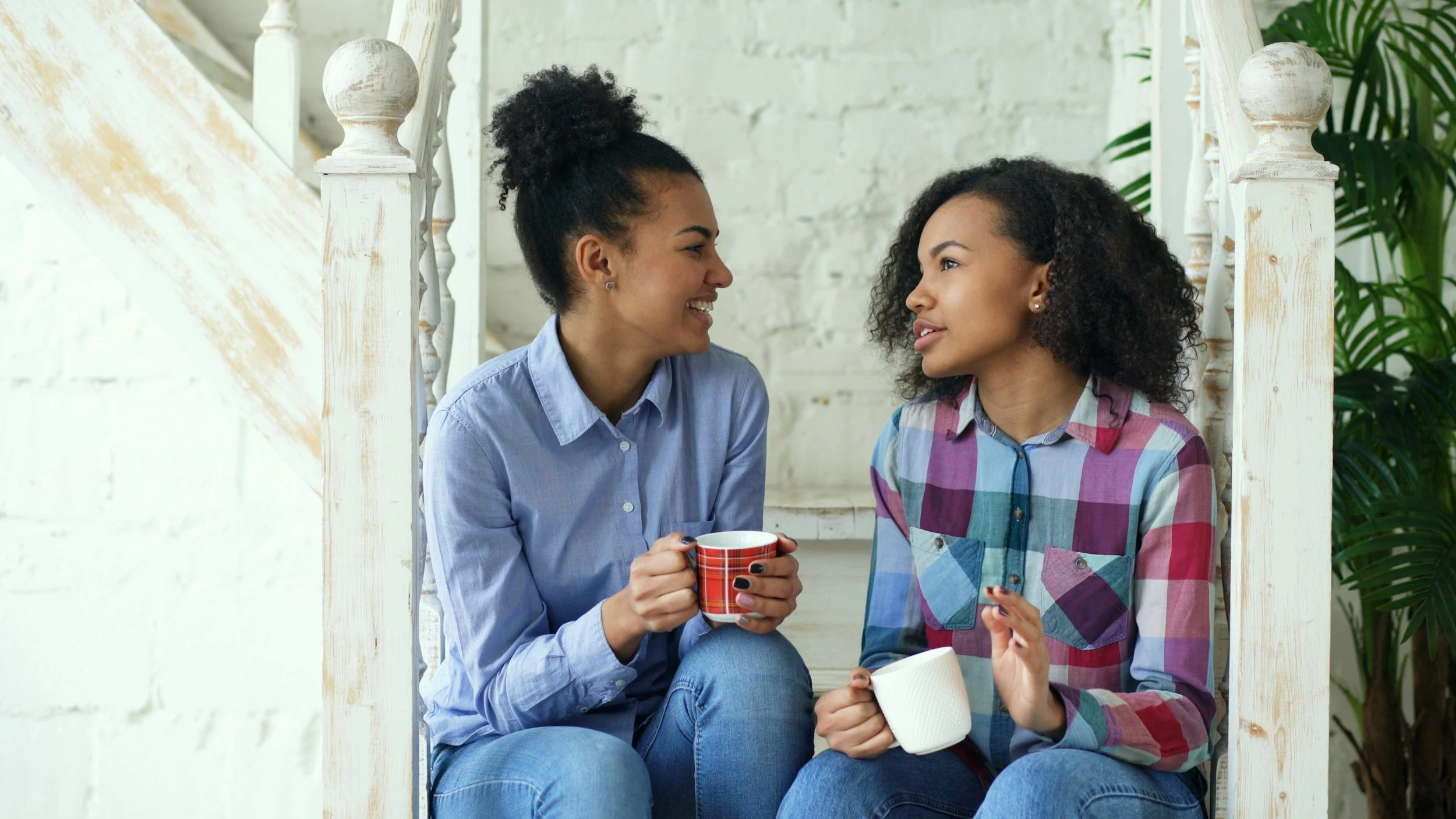 Two women sitting together on stairs, smiling and talking, each holding a mug, in a bright room with white brick walls and green plant in background.