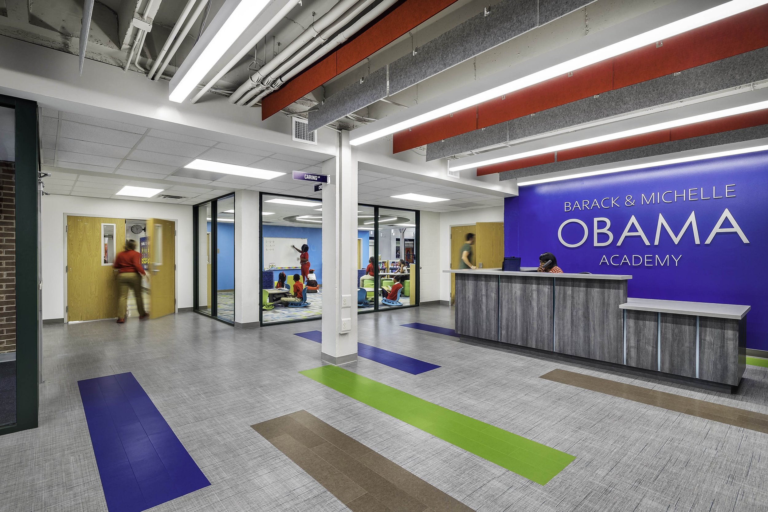 Interior of a school reception area with a front desk labeled "Barack & Michelle Obama Academy," bright blue wall, and an adjacent classroom visible through glass windows with children inside.