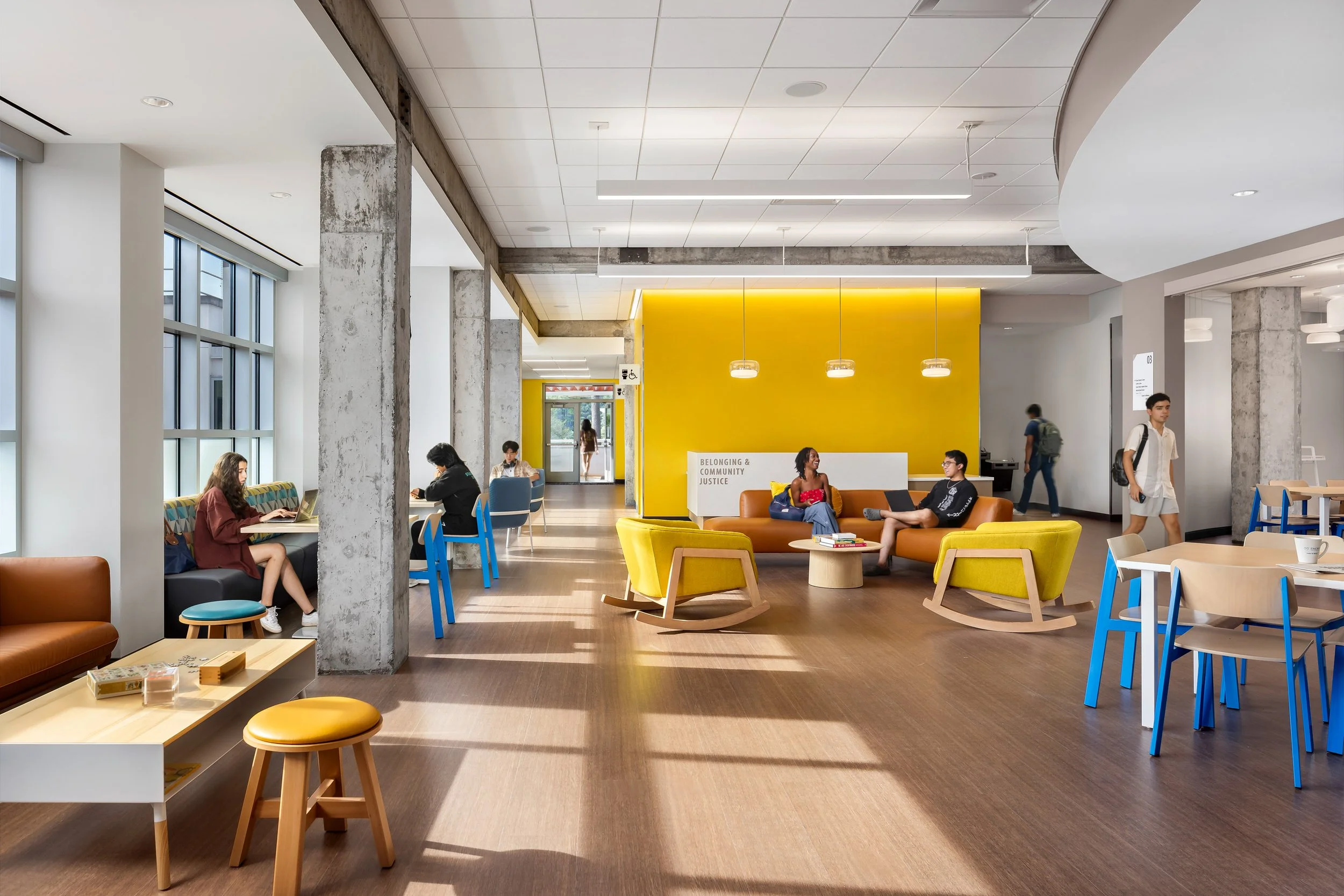 Modern college common area with students, featuring colorful furniture, large windows, and a yellow accent wall, promoting community and learning.