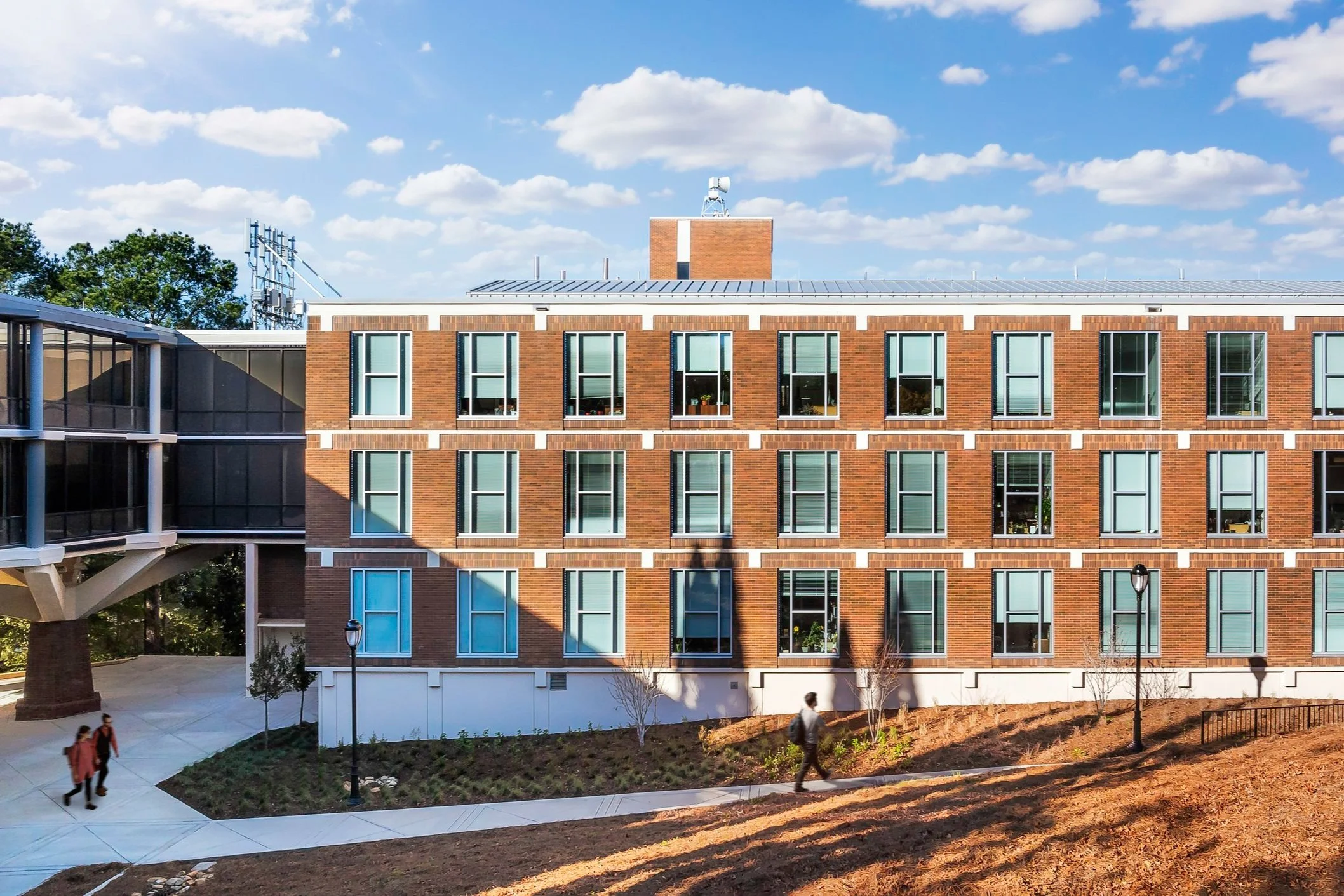 Brick academic building with modern windows under a blue sky with clouds, featuring a walkway and students.