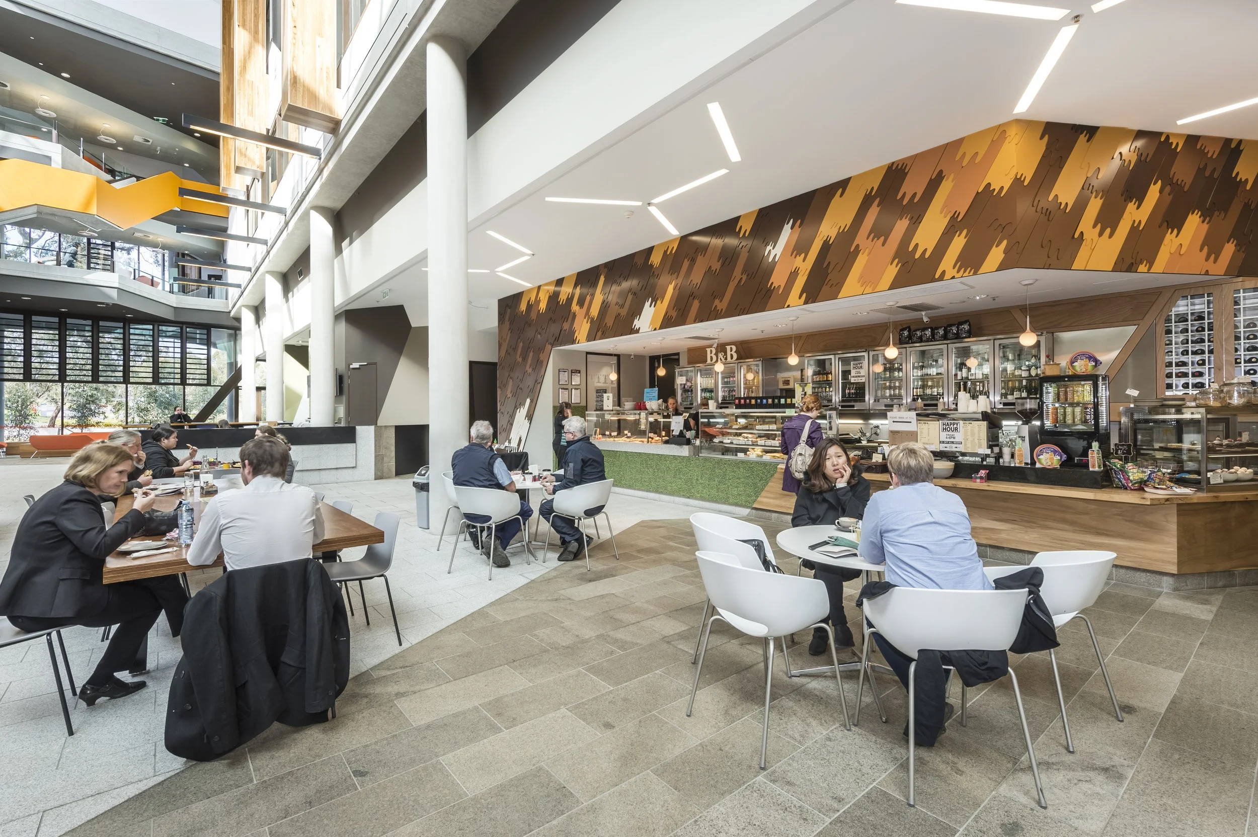 Interior of a modern coffee shop with multiple tables and customers, a counter with a variety of baked goods, and contemporary architectural design including large windows, white pillars, and decorative ceiling lighting.