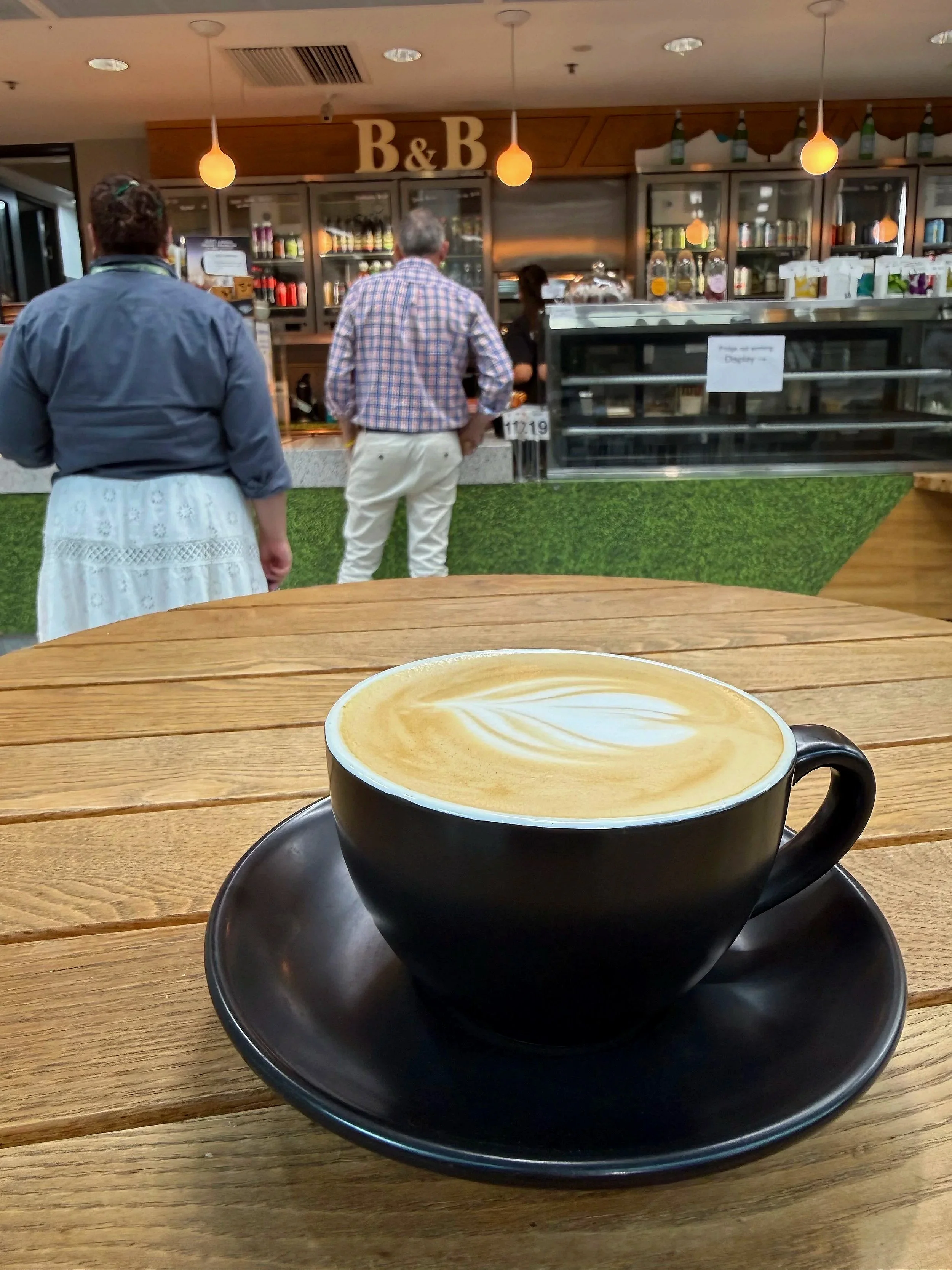 A black coffee cup with latte art on top, sitting on a black saucer on a wooden table inside a cafe. In the background, people are standing at the counter ordering, with a