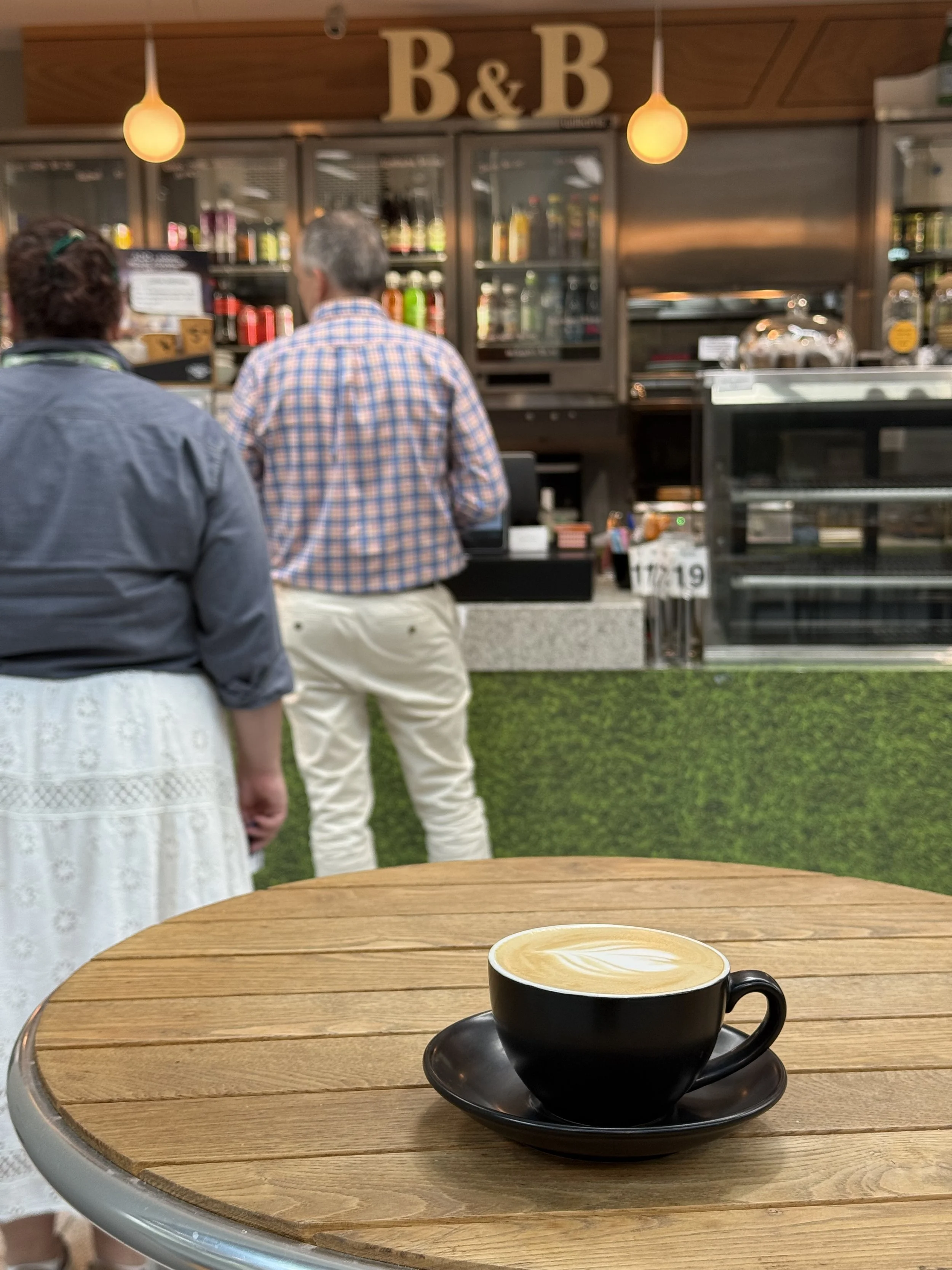 A black coffee cup with latte art on a wooden table in a cafe, with two people standing at the counter in the background.