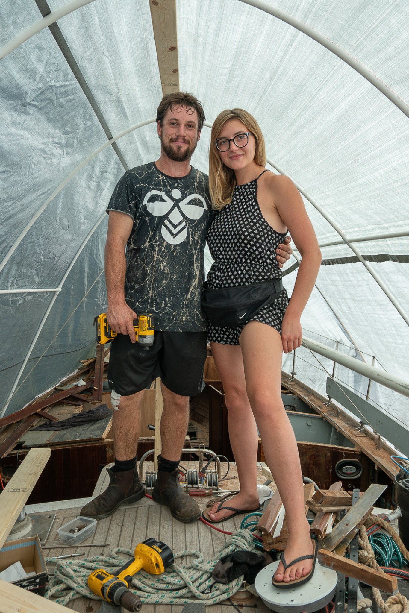 A man and woman standing inside a boat under construction, smiling at the camera. The man has a beard, wears a graphic t-shirt, shorts, and holds a power drill. The woman has glasses, blonde hair, wears a tank top, shorts, flip flops, and has a waist