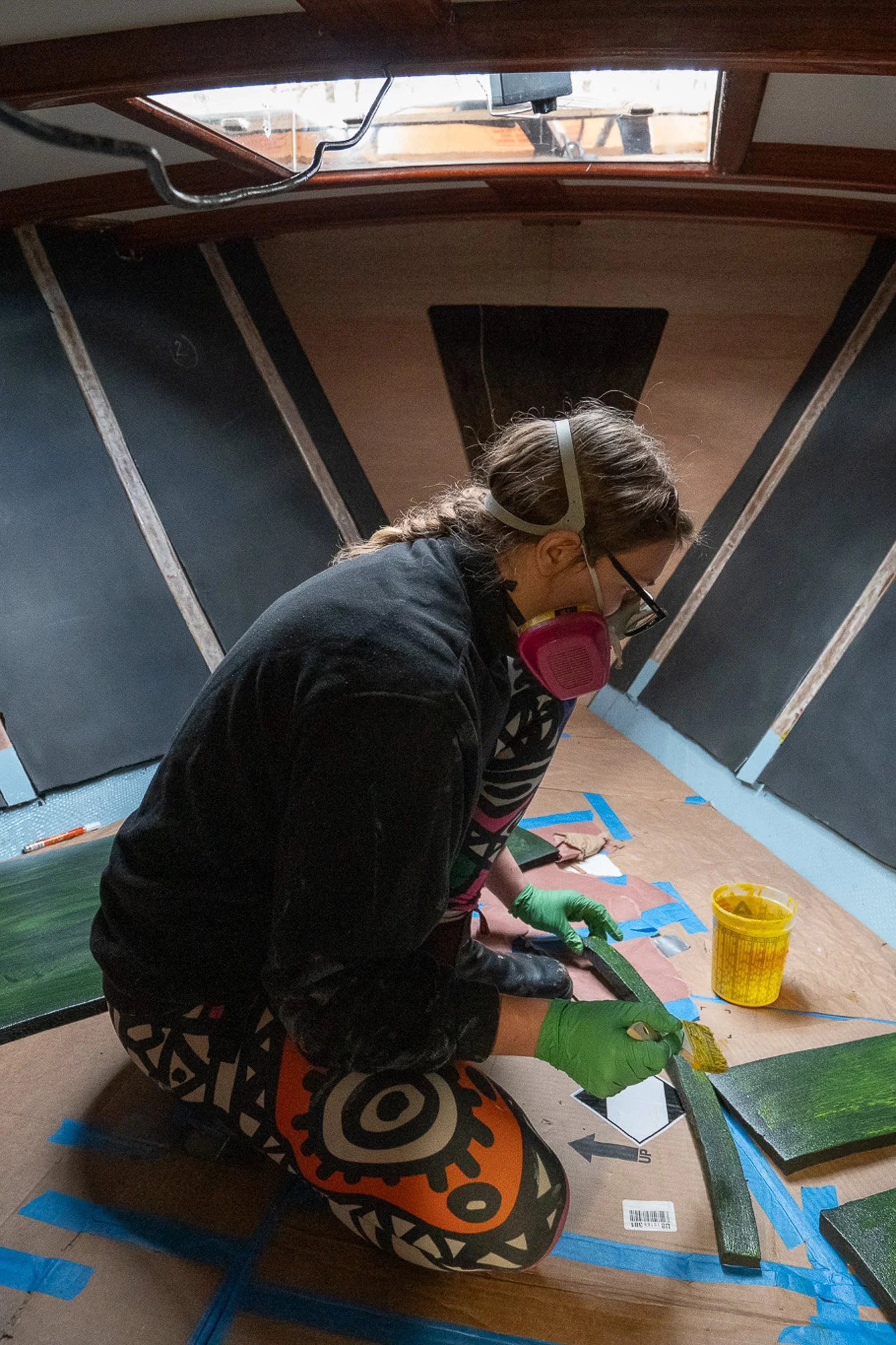 Person kneeling on a floor, working on a project with a paintbrush, wearing green gloves, a mask, glasses, and gear patterned pants, in a room with black walls and a wooden ceiling.