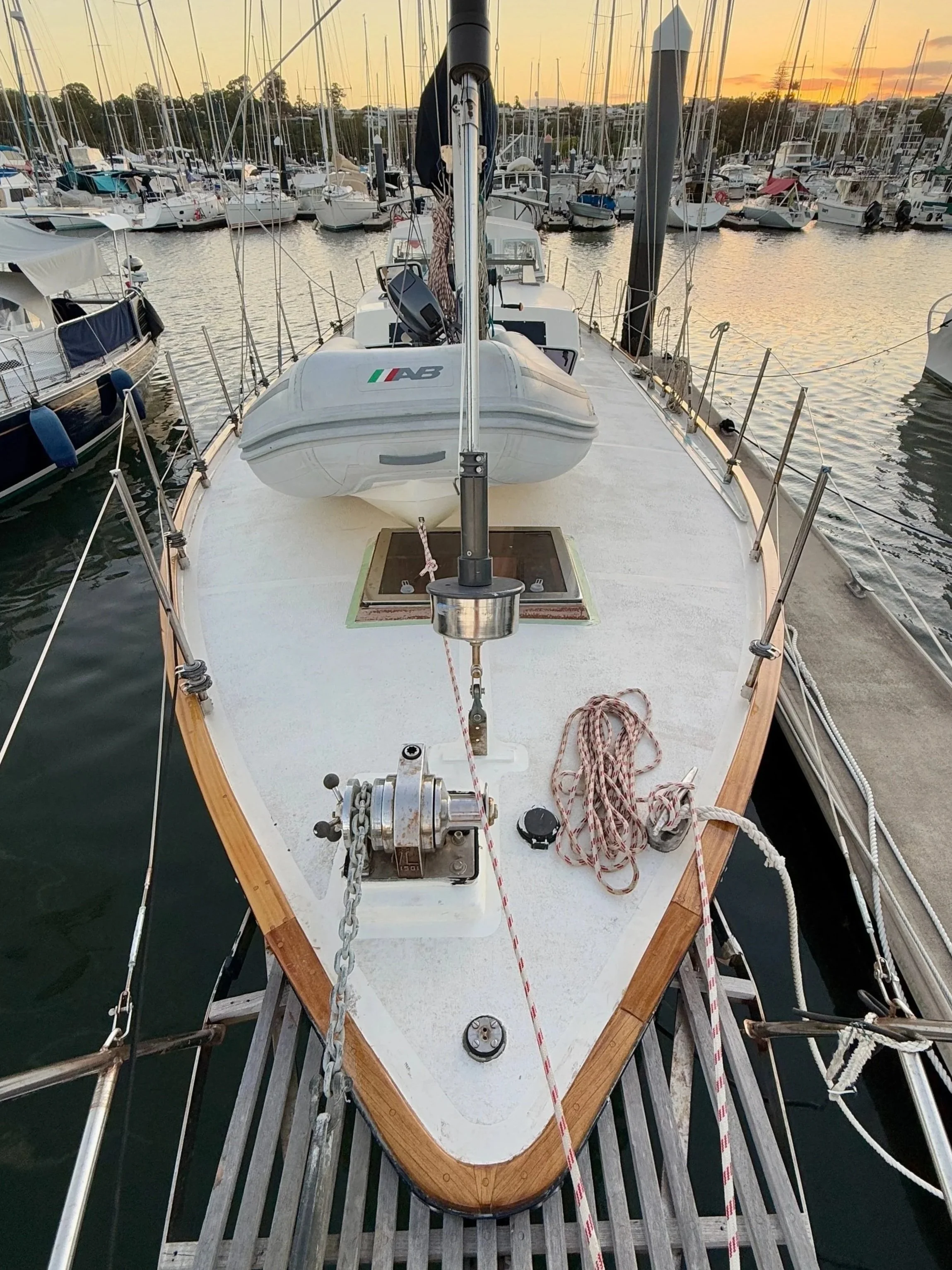 Front view of a sailboat docked at a marina during sunset, showing the deck, ropes, and surrounding boats.