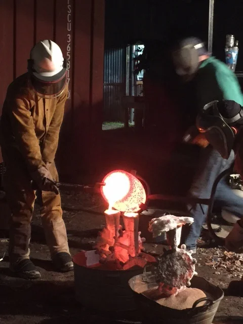 Carlos and Will lift the hot crucible of hot bronze out of the furnace and pour it into the shells. The one holding the pour end is called the dead men.