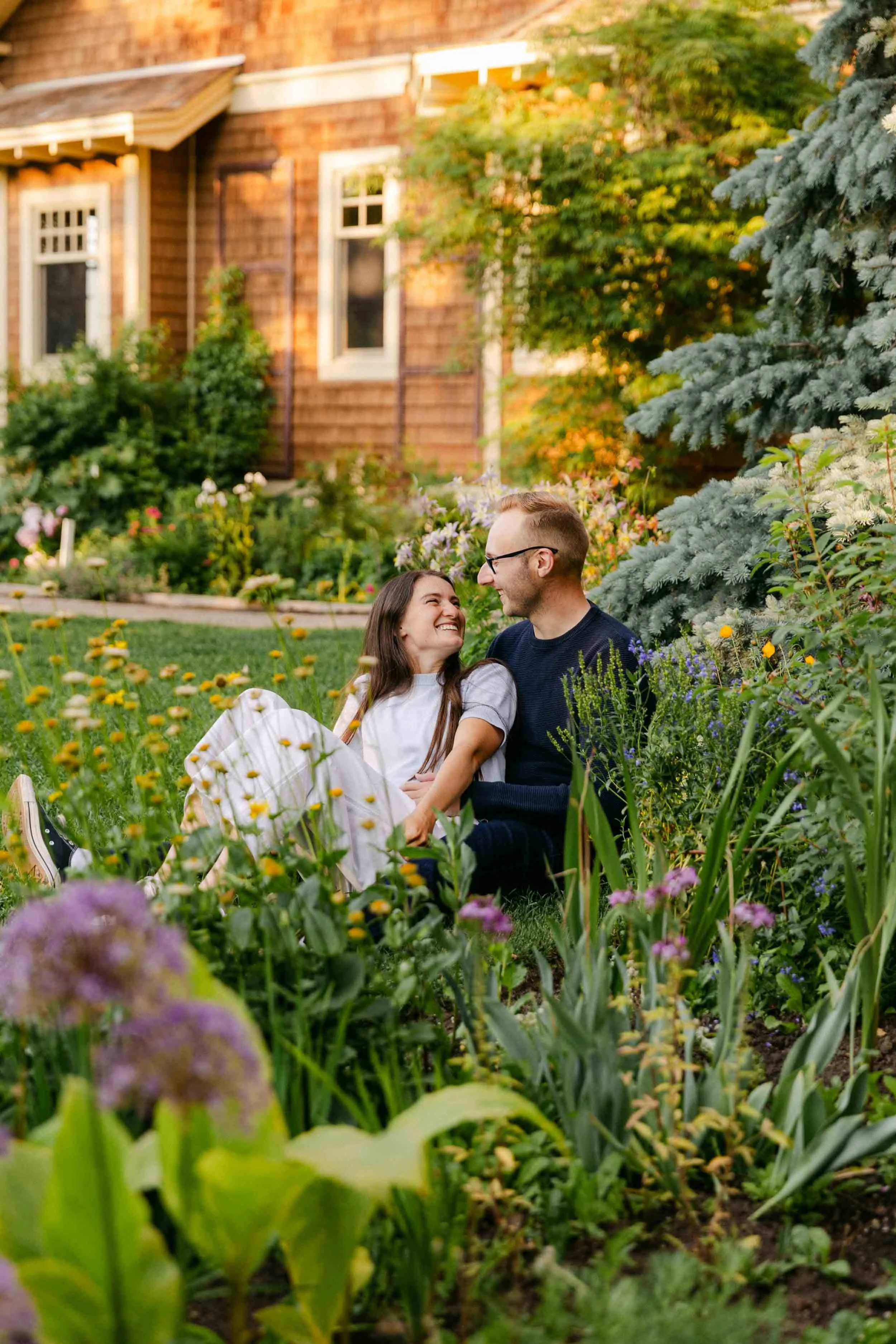 A couples engagement photo session at Reader Rock Garden in Calgary, wearing shades of blue in front of blooming yellow and purple flowers with while they gaze at one another lovingly