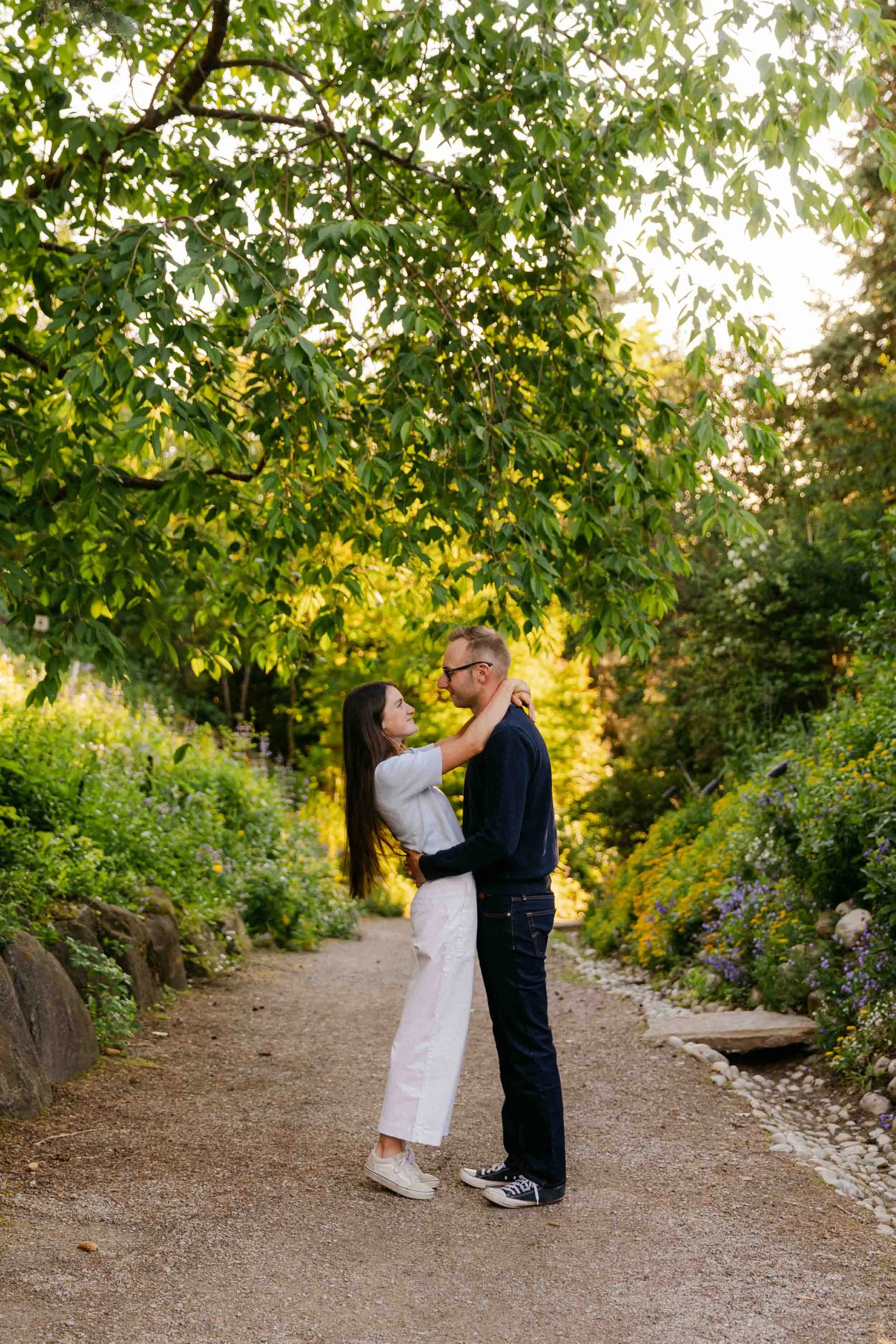 A couples engagement photo session at Reader Rock Garden in Calgary, wearing shades of blue with white pants on a gravel path with trees overhead while he pulls her close