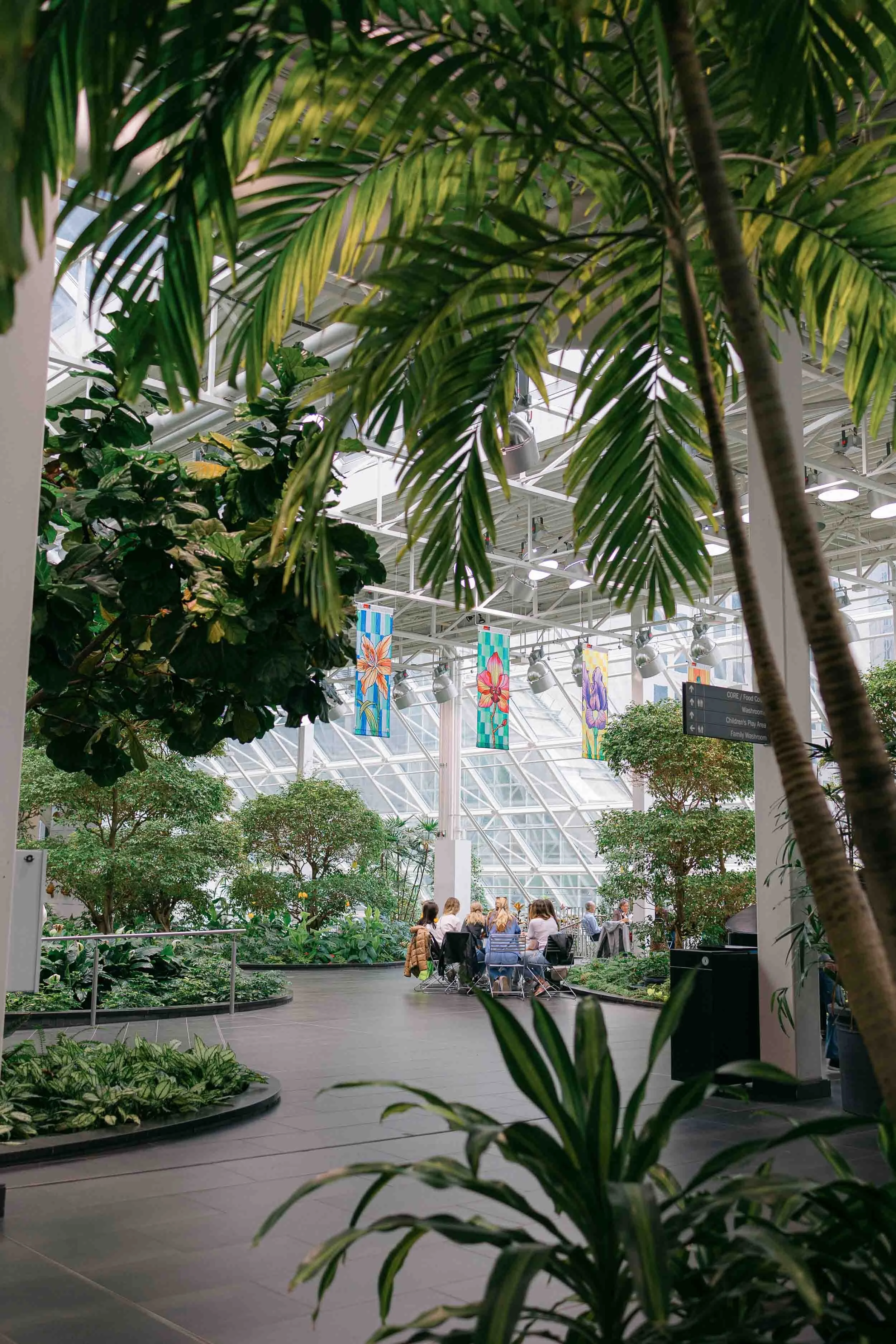 Inside Calgary's indoor Devonian Gardens, a gathering of women under iris banners, surrounded by tropical greenery and curved pathways