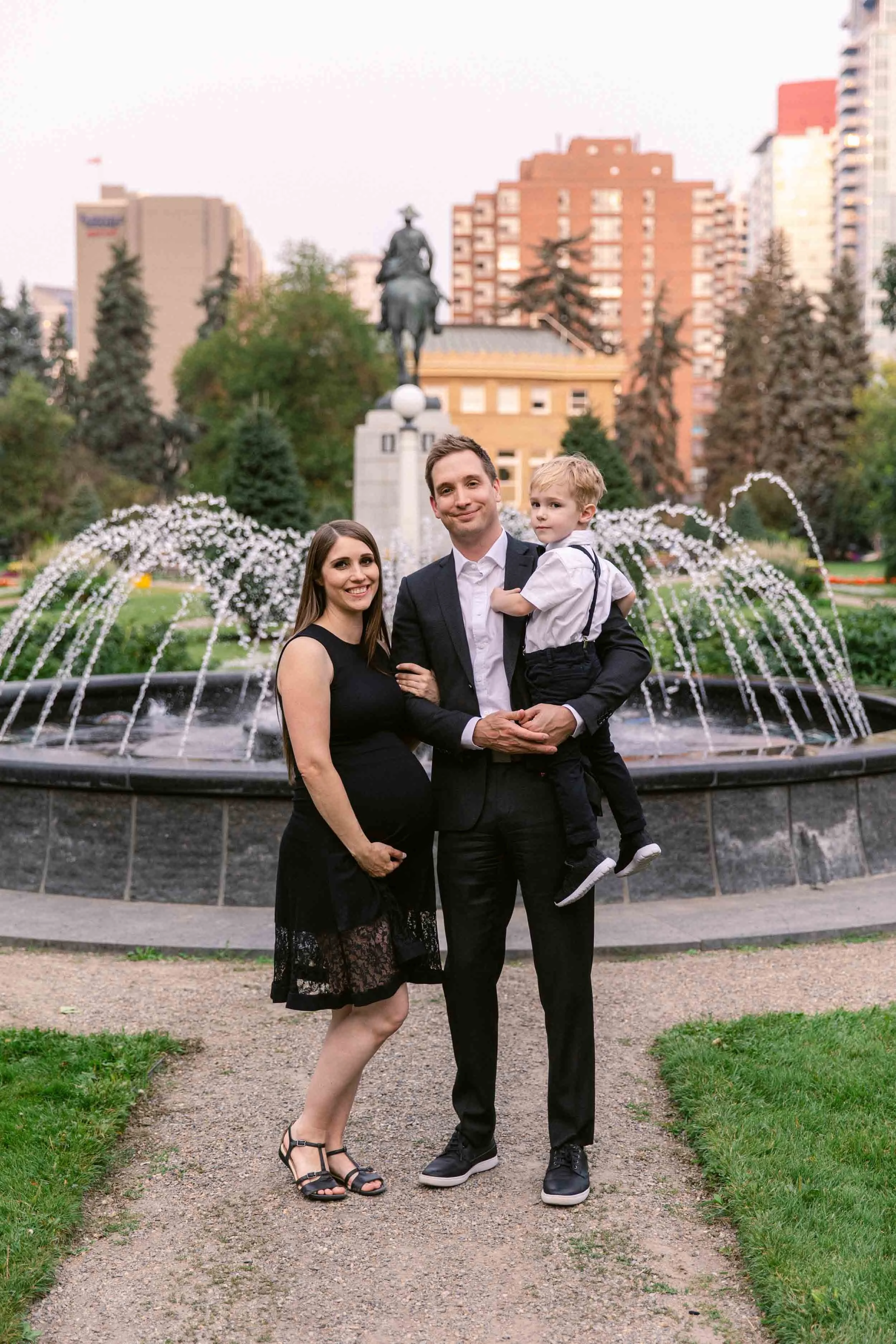 Maternity photo session at Central memorial garden in calgary, a family dressed up formally in black suit, suspenders and lace dress, in front of water fountain