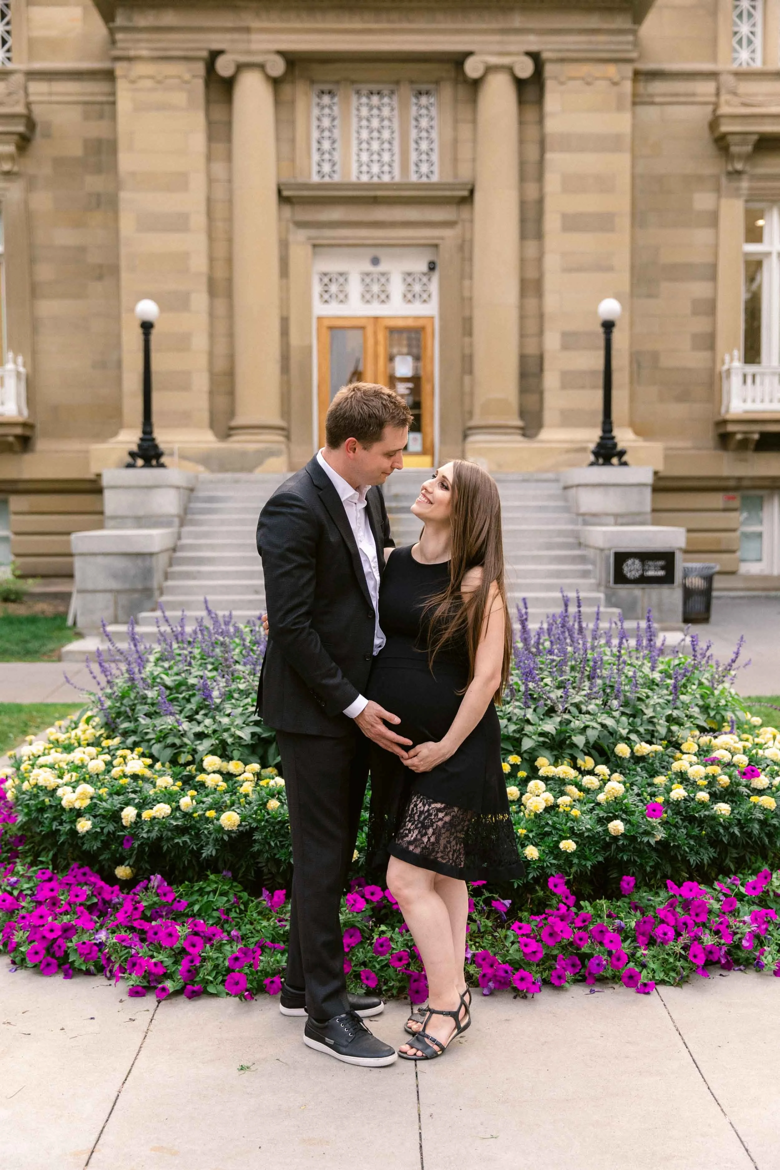 Maternity photo session at Central memorial garden in calgary, a family dressed up formally in black suit, suspenders and lace dress, in front of classical architecture, the oldest library in the city