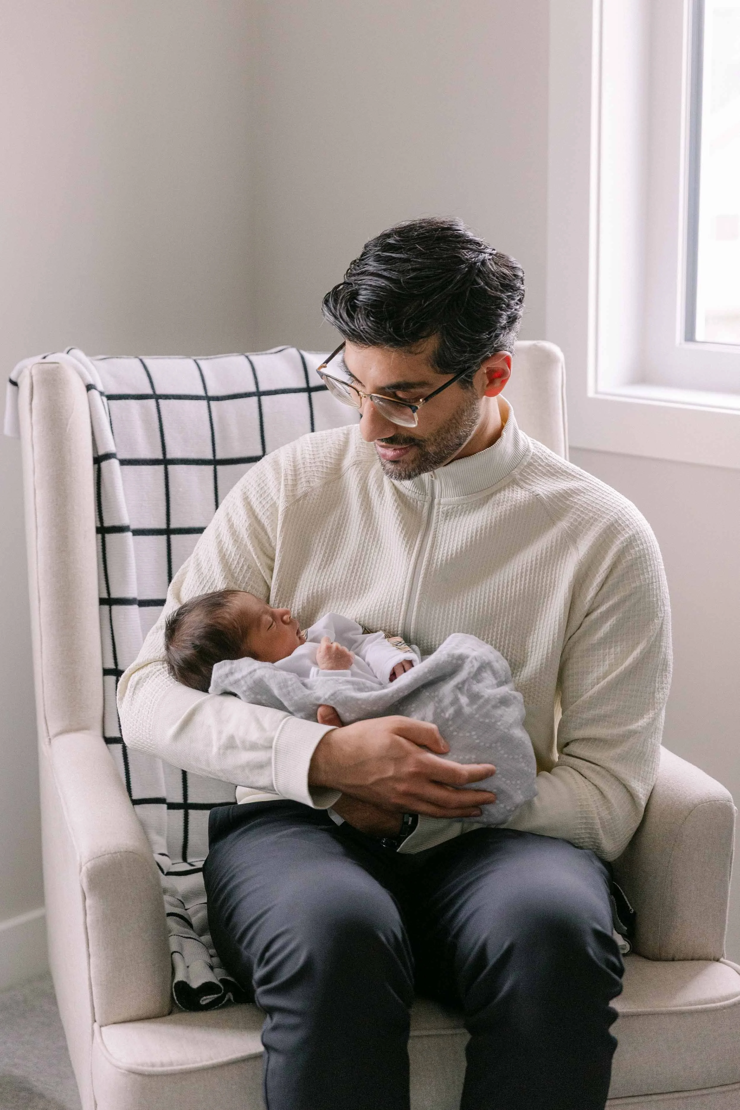 dad and baby in upholstered cream rocking chair, newborn lifestyle shoot, wearing cream, white and black neutrals, indian father and son