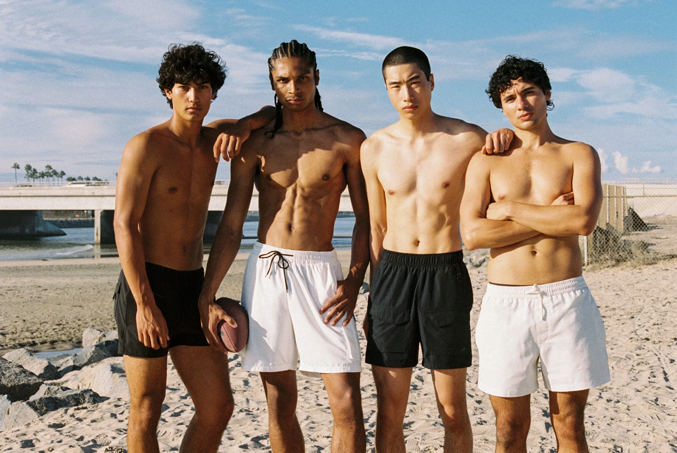 Four shirtless young men standing together on the beach with arms around each other's shoulders, looking at the camera, with a blue sky and a bridge in the background.