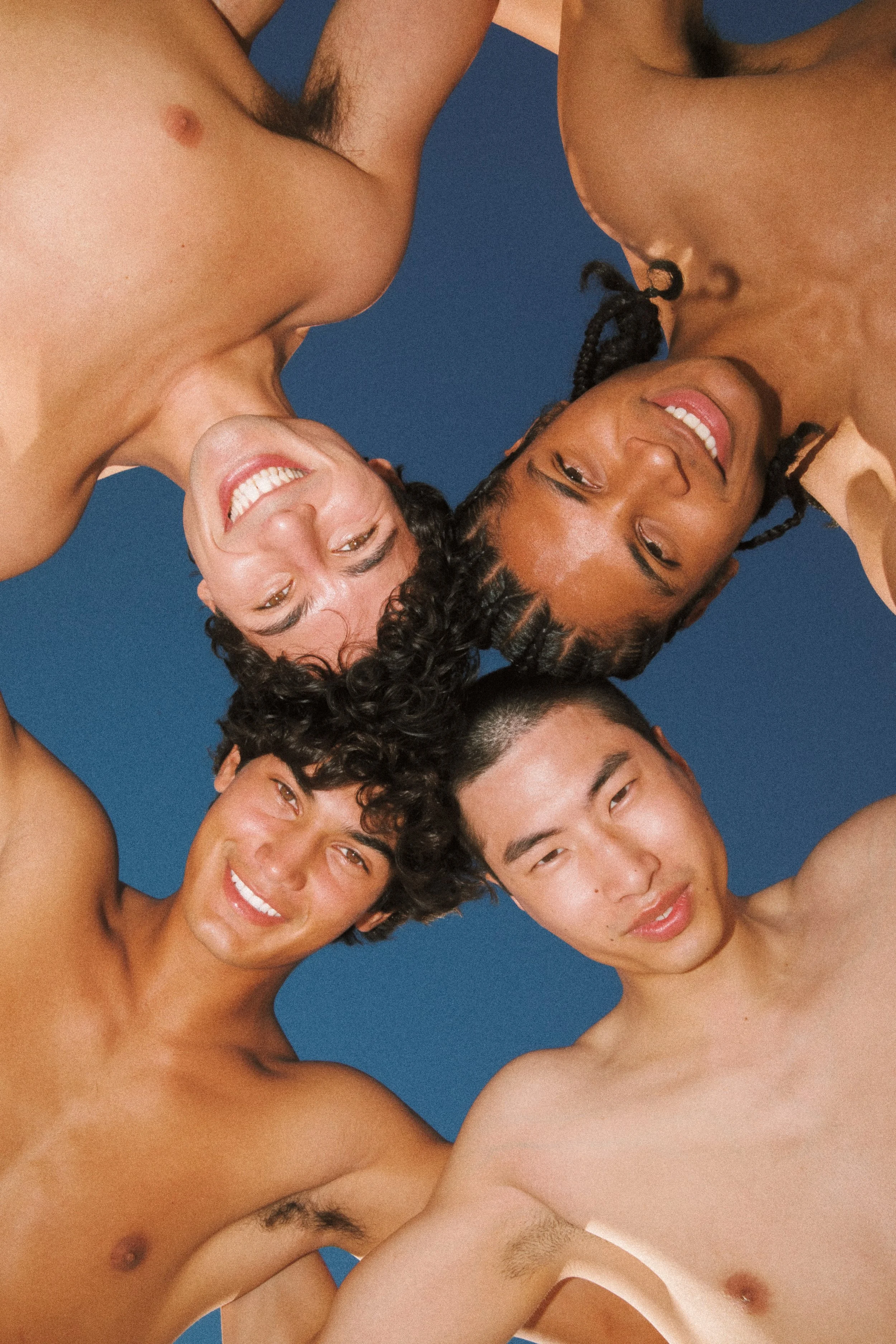 Group of topless diverse young adults, smiling and leaning toward the camera against a blue background.