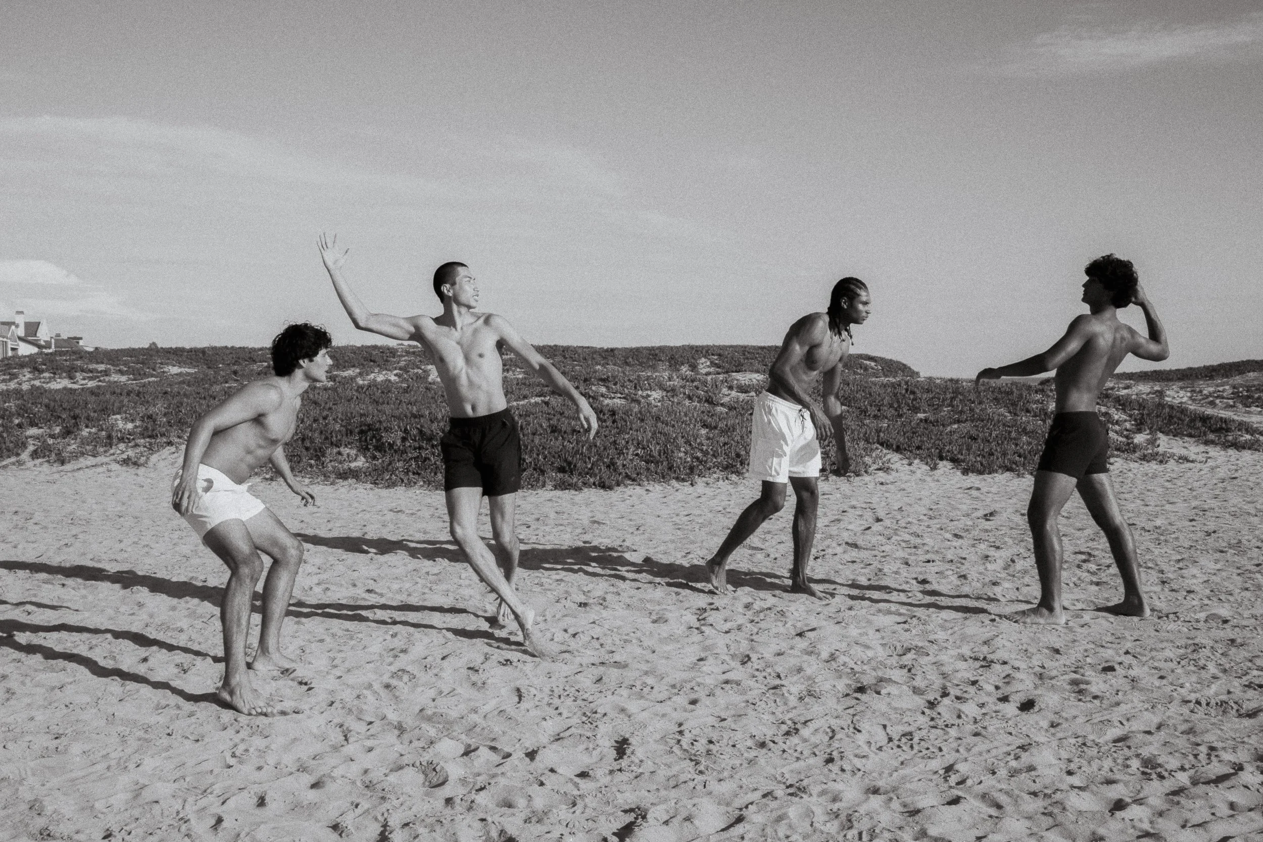 Black and white photo of four young men talking on a sandy beach with dunes and buildings in the background.