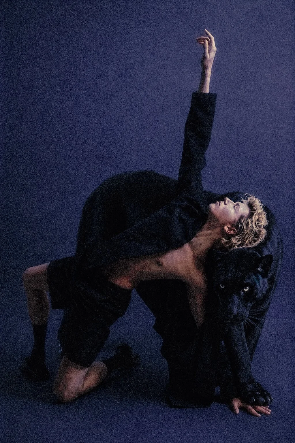 Person with curly blonde hair wearing black attire, positioned on all fours, with one arm extended upward, alongside a black panther against a dark background.