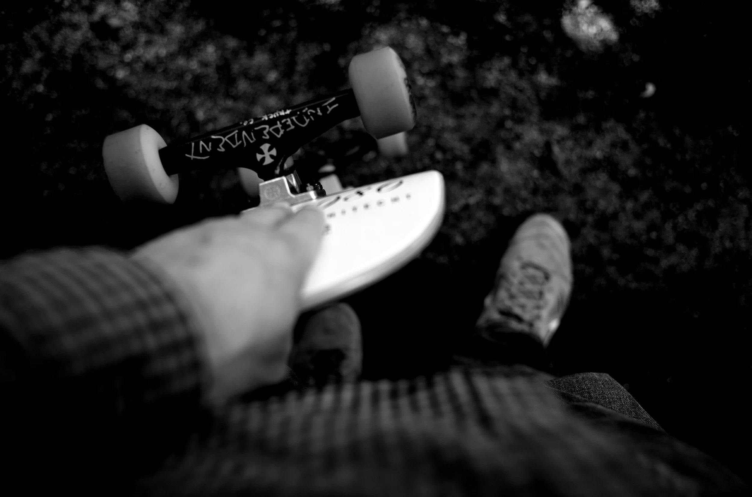 A person holding a skateboard with one hand, standing on a rough ground surface, with the tip of a shoe visible on the pavement.