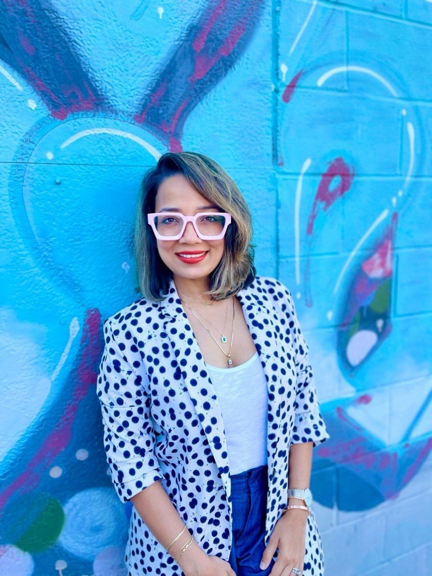 A person in a polka dot jacket and pink glasses standing in front of a blue wall Description automatically generated.jpeg