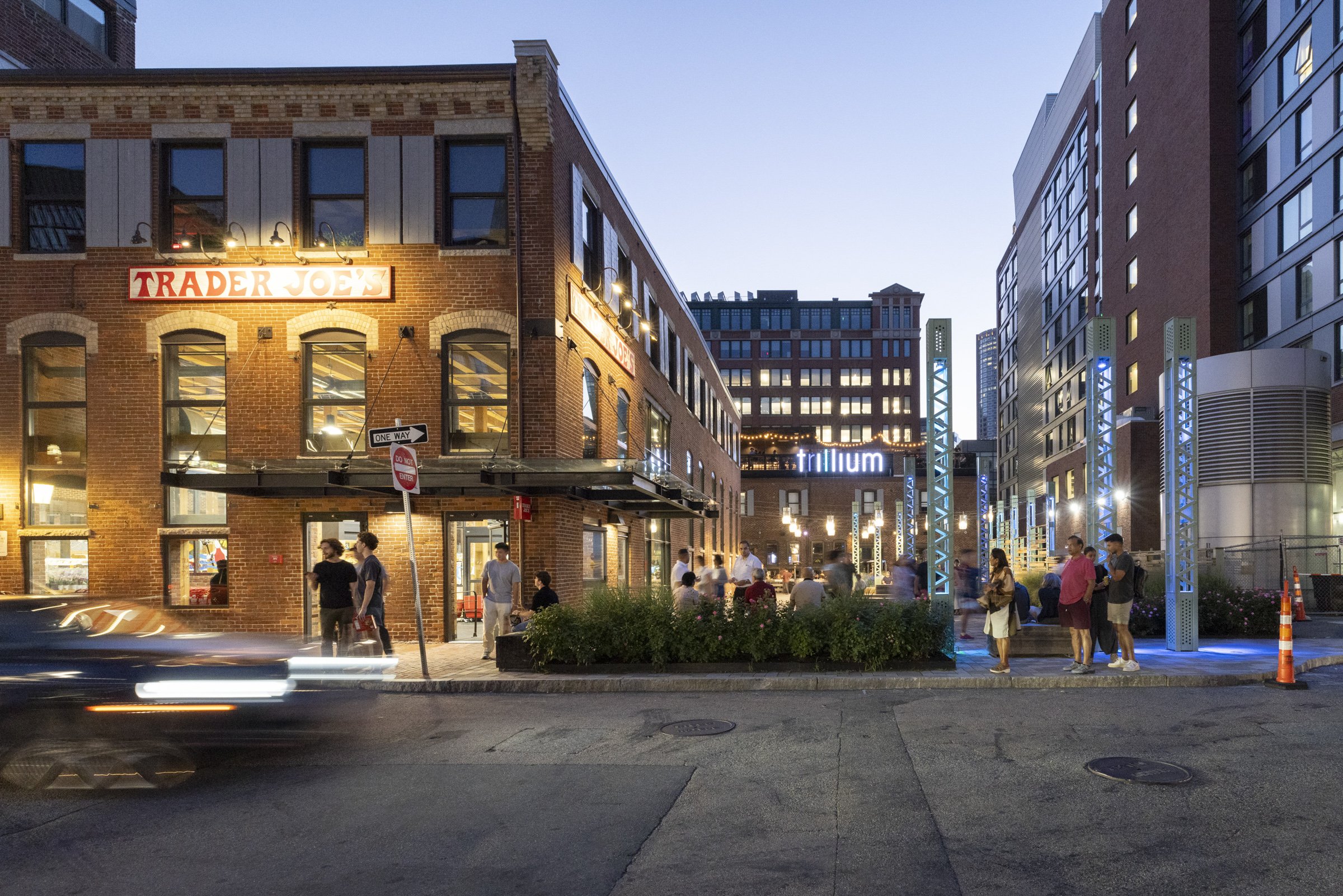 A busy city street scene at dusk with a brick building on the left featuring a Trader Joe's sign, and a group of people gathered along a sidewalk with decorative blue-lit structures and modern buildings in the background.