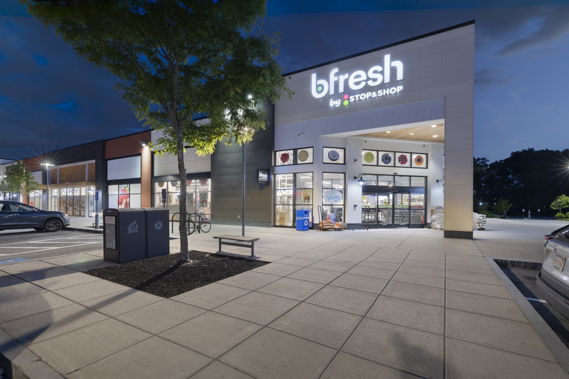 Night view of the exterior of a modern grocery store called "b.fresh by STOP & SHOP" with a lit sign, an entrance with glass doors, a bench, trees, a trash bin, and parking spaces, some cars, and a bicycle nearby.