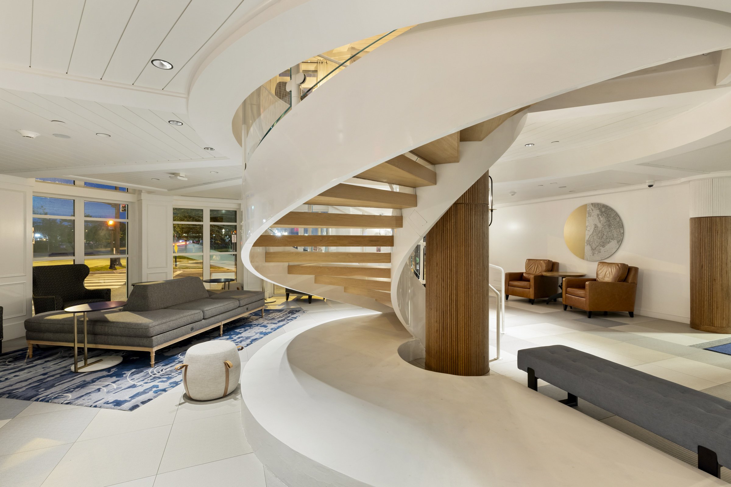 Modern hotel lobby with a curved staircase, gray sofa, armchairs, and decorative wall art.