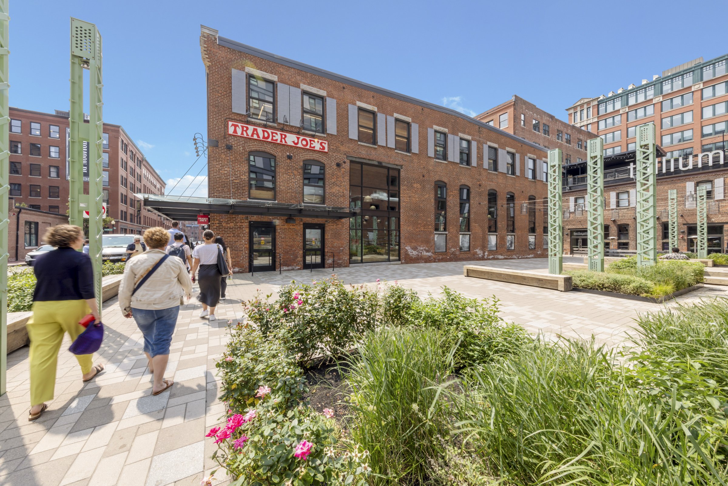 People walking on a brick sidewalk in front of a red brick Trader Joe's store, with greenery and flowering bushes in the foreground and modern buildings nearby.