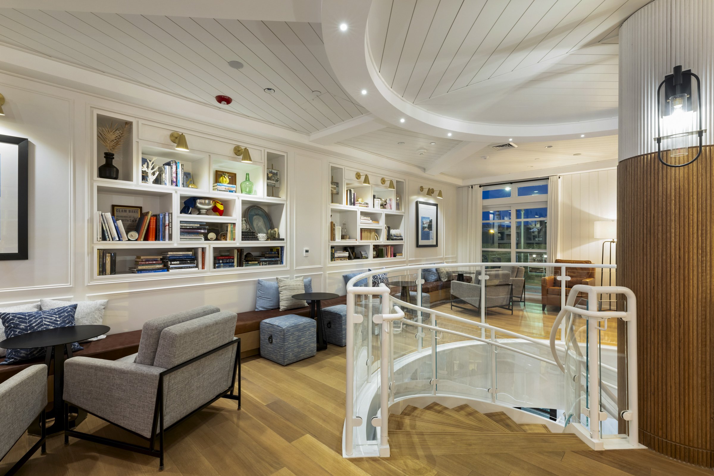 A cozy lounge area with built-in white bookshelves filled with books and decorative items, a staircase with a white modern railing, multiple seating areas with armchairs and sofas, wooden floors, a large window with white curtains, and contemporary l