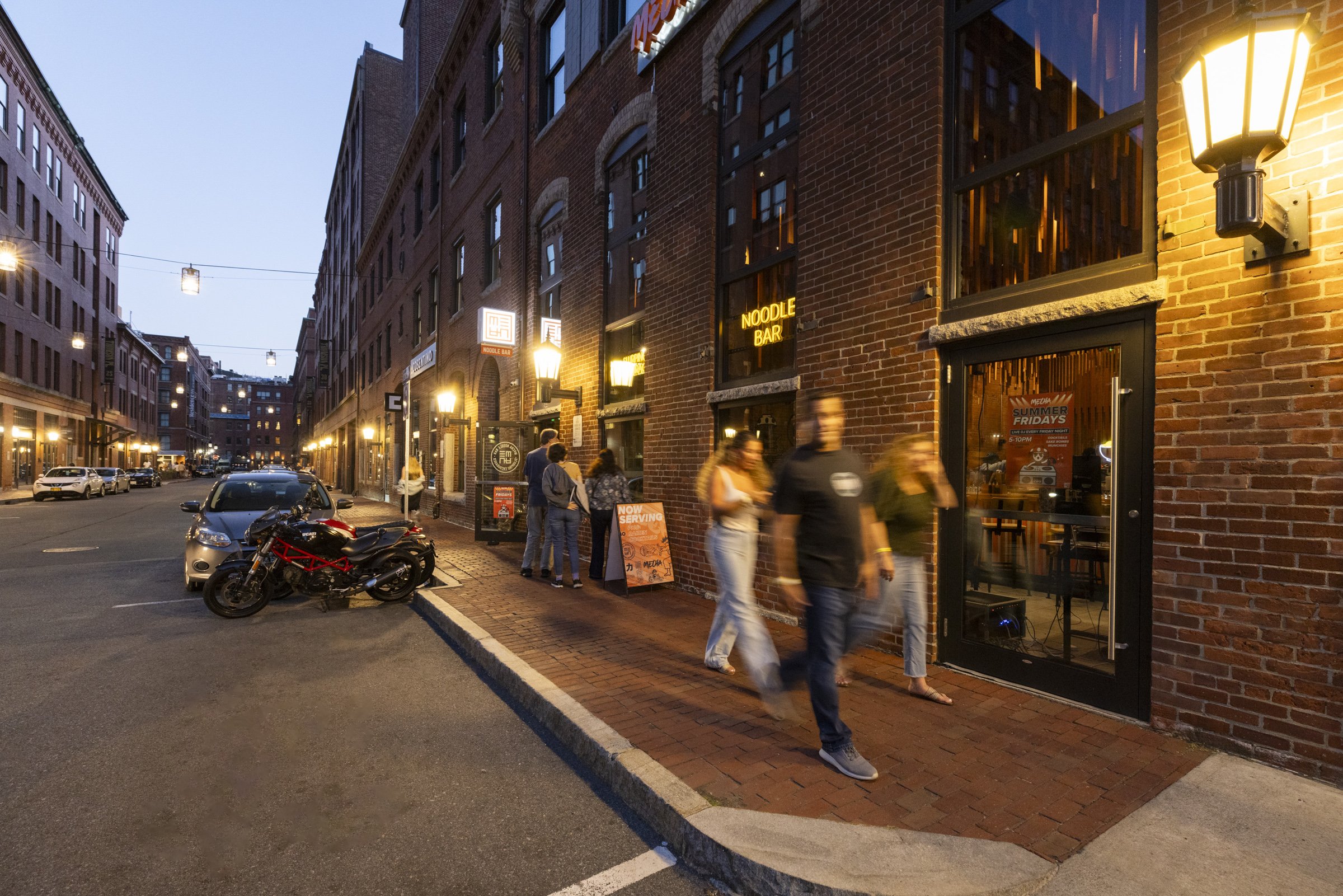 People walking past a brick building with illuminated signs that say 'Noodle Bar' and 'SUMMER FRIDAYS,' on a city street at dusk, with parked cars and motorcycles.