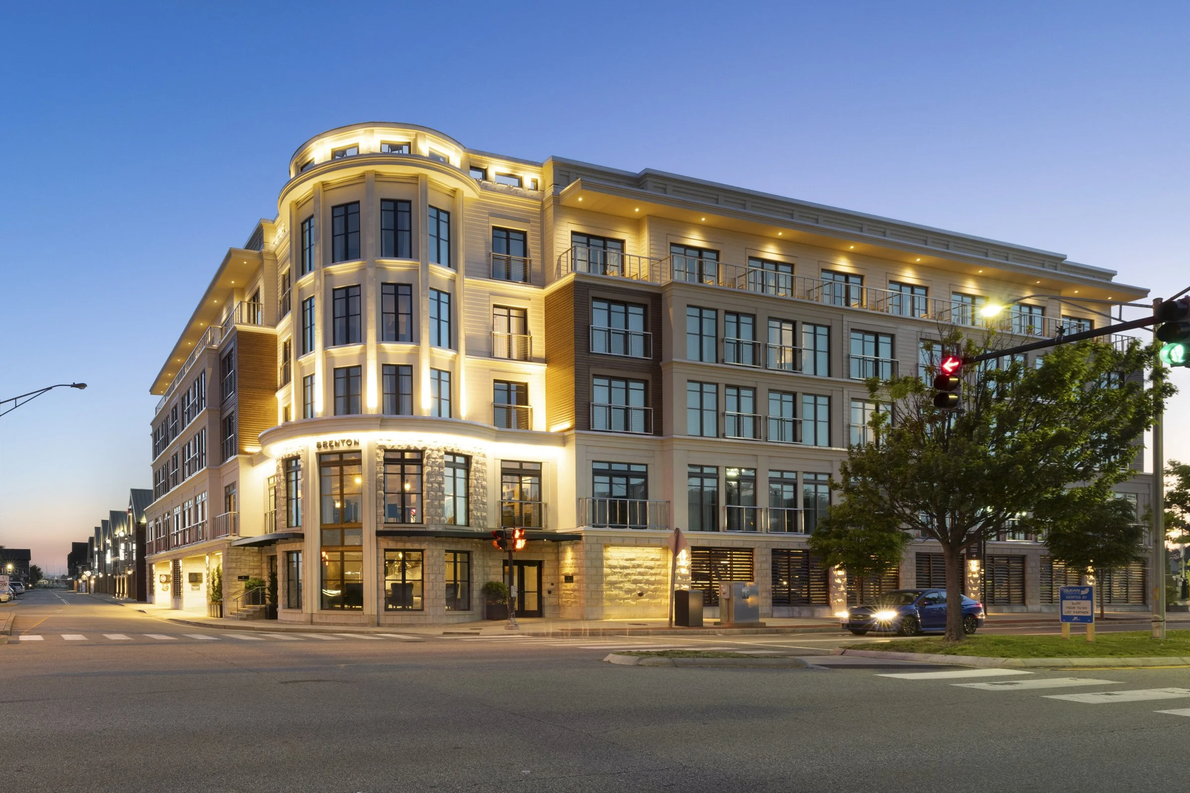 Modern multi-story apartment or hotel building with illuminated exterior at dusk, corner location, with a tree, traffic lights, and parked cars nearby.