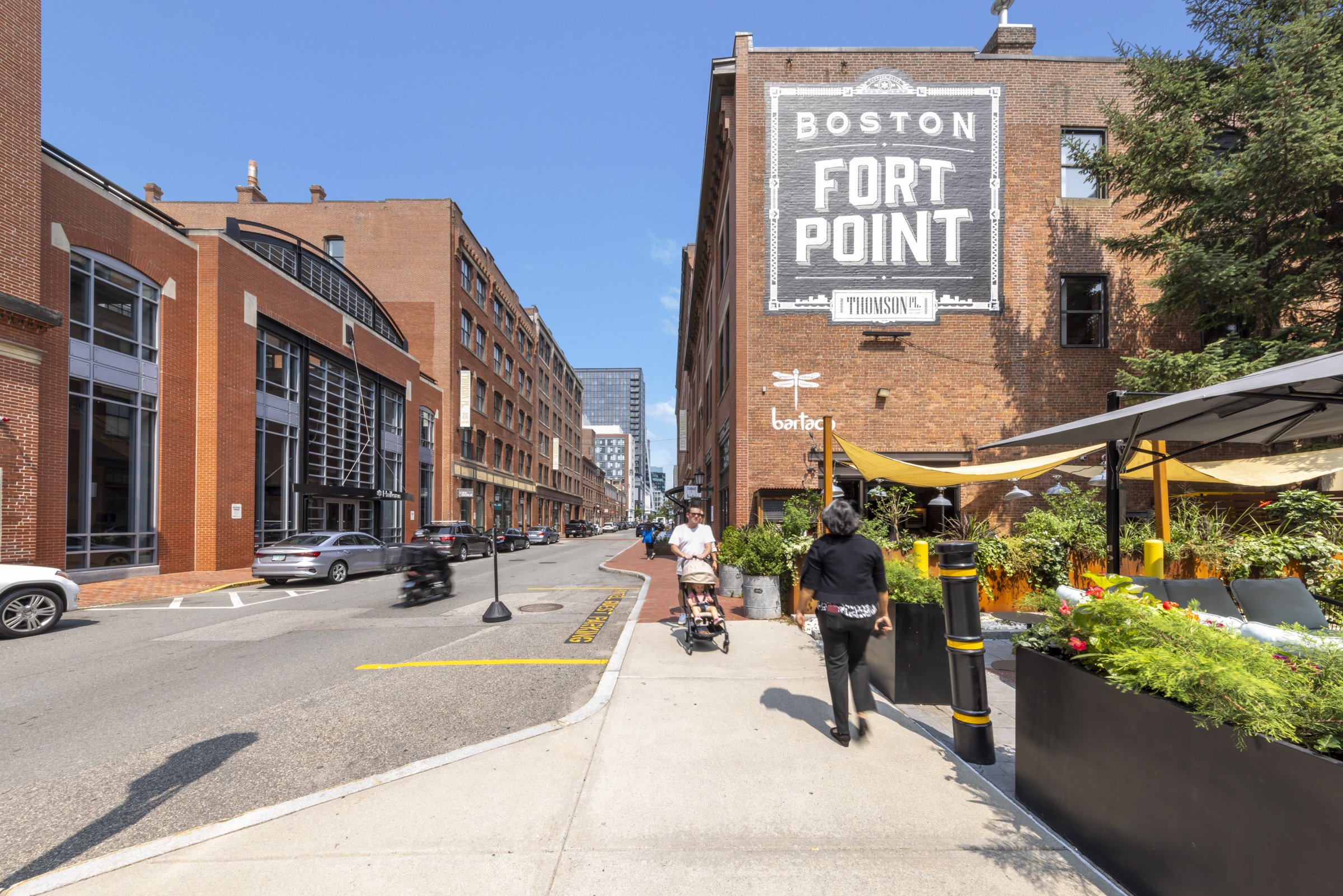 Street view of Fort Point in Boston with people walking, cars parked along the curb, brick buildings, and a large sign on the building reading 'Fort Point Boston'.