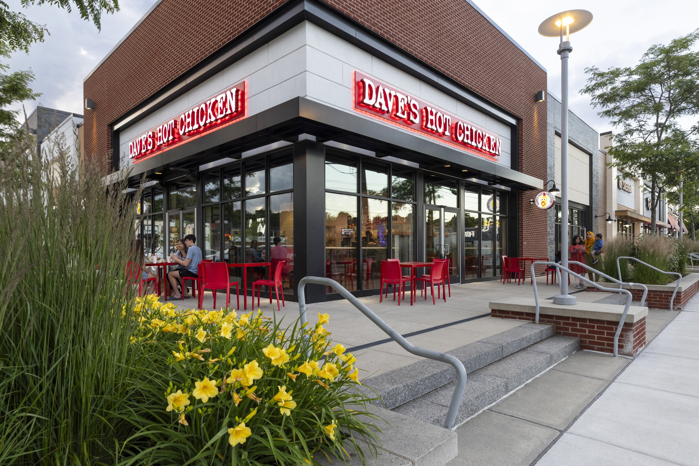 Exterior of a restaurant with red chairs on the patio, large glass windows, and a sign that reads 'DAVE'S HOT CHICKEN' in red neon lights, with a girl and a woman walking nearby.