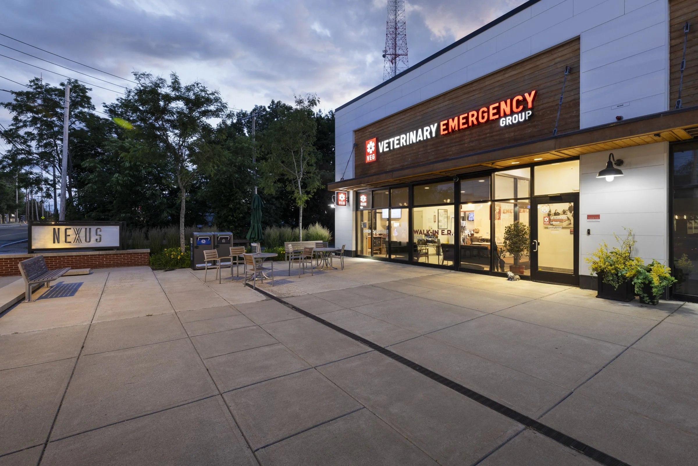 Exterior view of a veterinary emergency clinic during evening hours with outdoor seating, greenery, and illuminated signage.