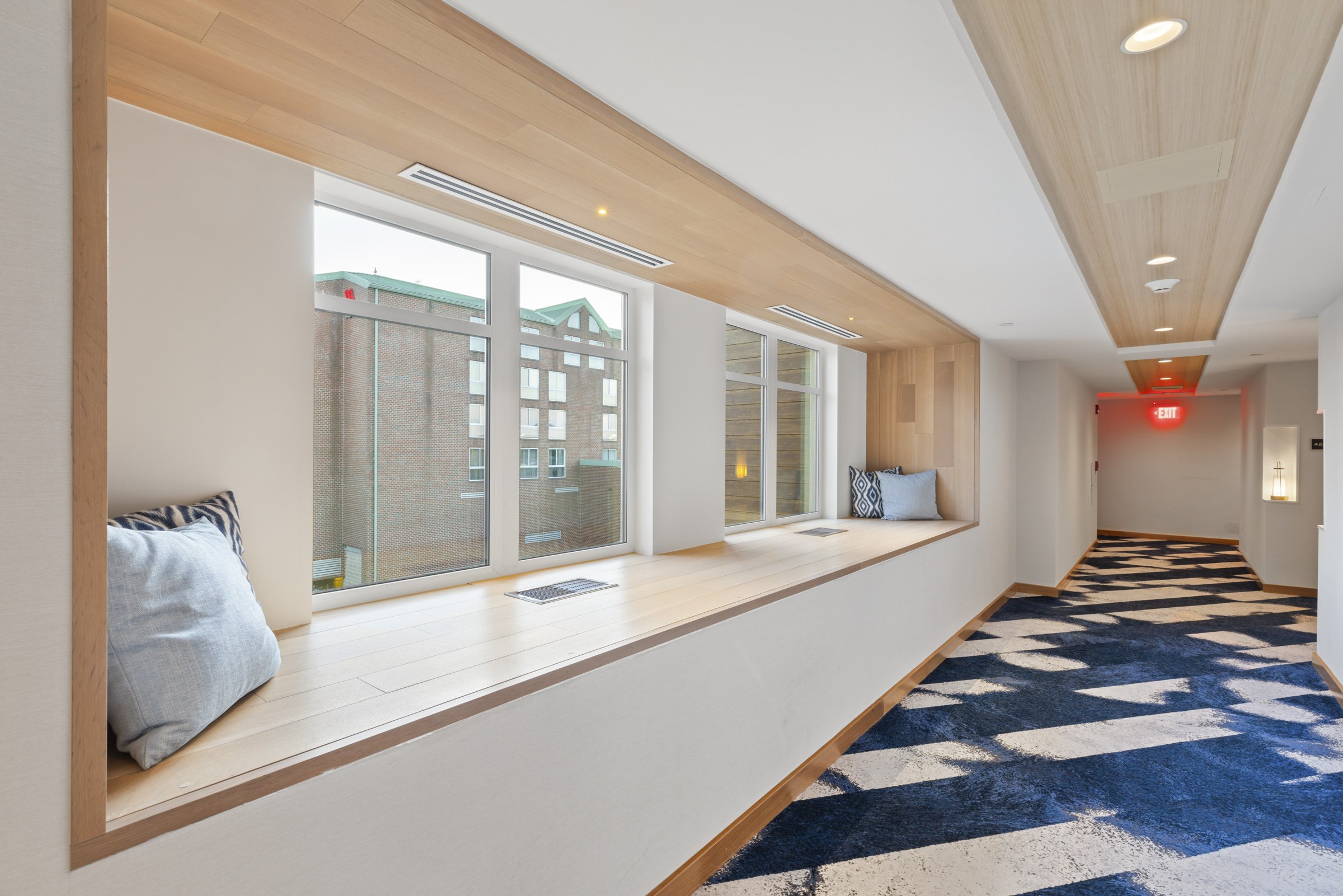 Contemporary corridor with large window seats with pillows, wooden accents on ceiling and window ledge, patterned blue and white carpet, and red exit sign at the end.