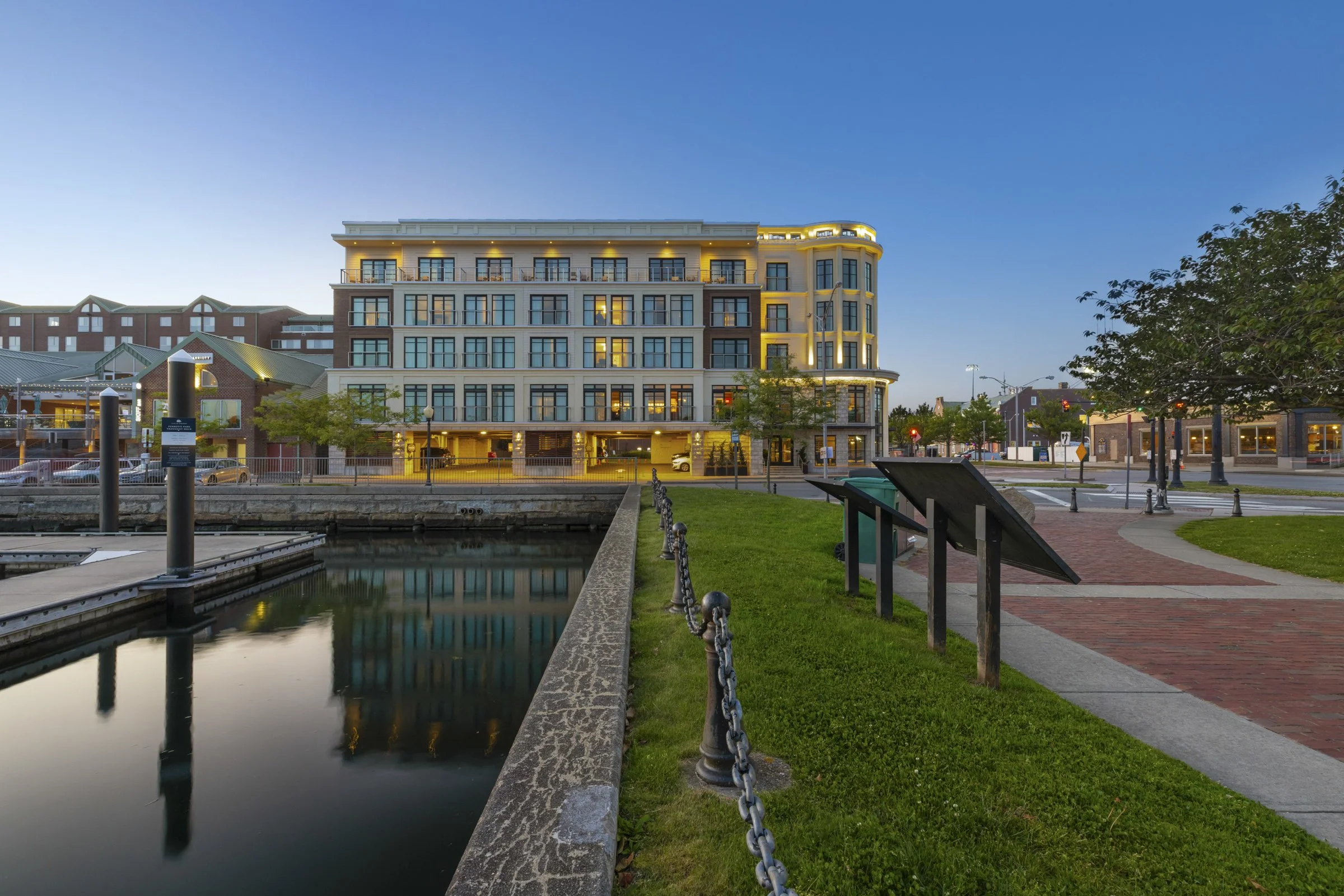 A scenic view of a waterfront sidewalk with a modern multi-story building, trees, street lamps, and a clear evening sky.